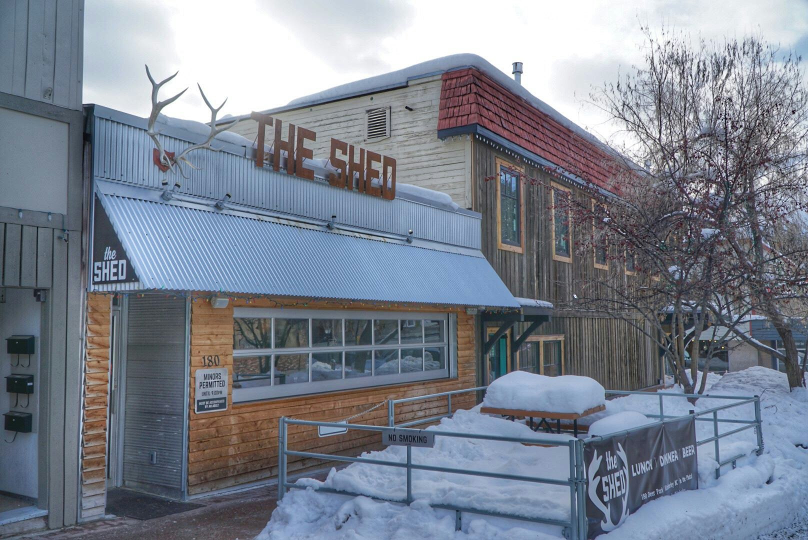 The exterior of a bar/restaurant, ; it has horizonal wood cladding and a corrugated metal awning decorated with a set of antlers and The Shed in red, cut-out letters. A few tables in an outdoor seating area are laden with several feet of snow.