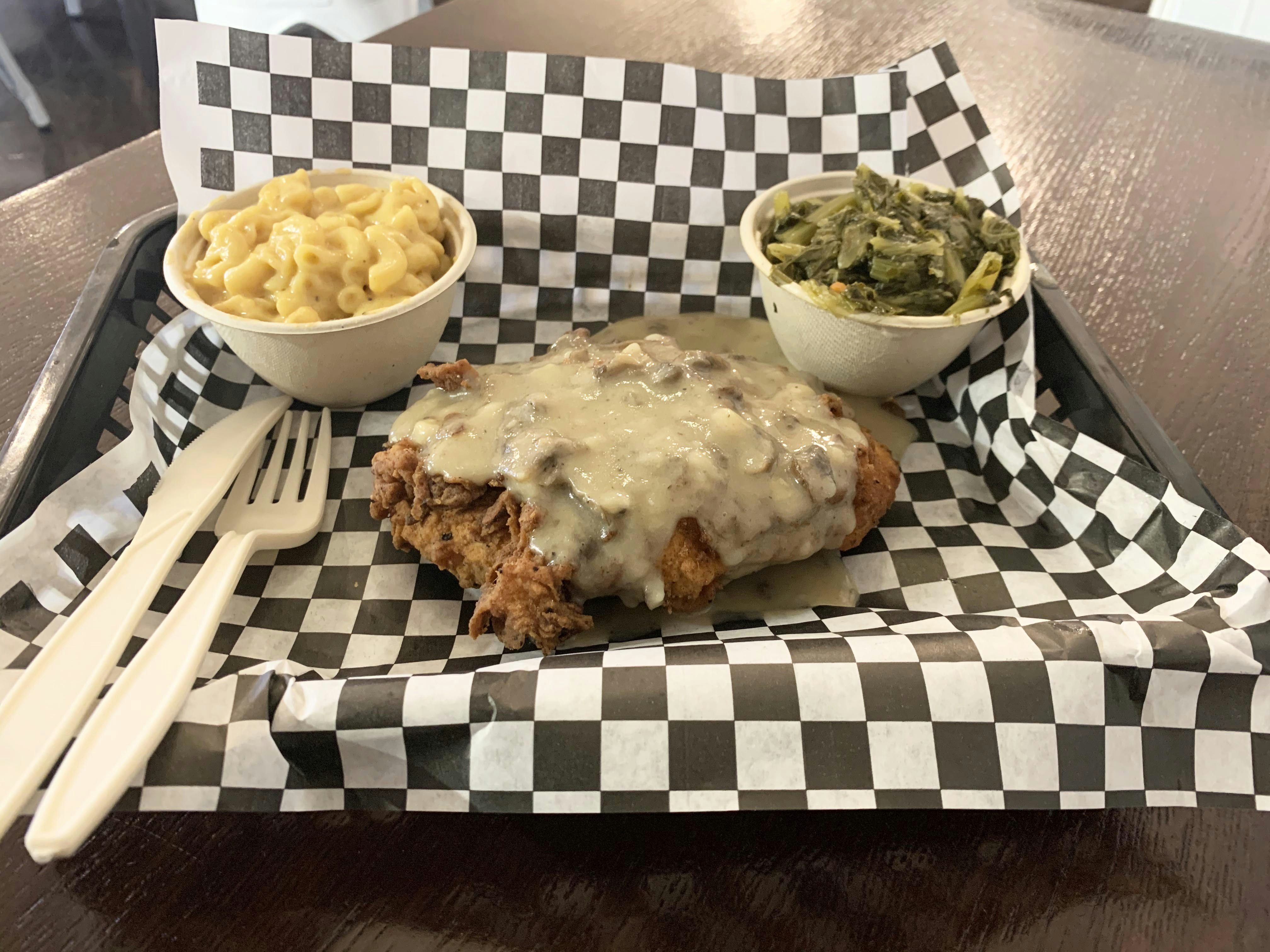 Small bowls of vegan mac and cheese, turnip greens sit on opposite sides of small plastic tray lined with black and white checkered paper. In the center is vegan fried chicken smothered in mushroom sauce. A plastic knife and fork rest on the left-hand side of the tray.