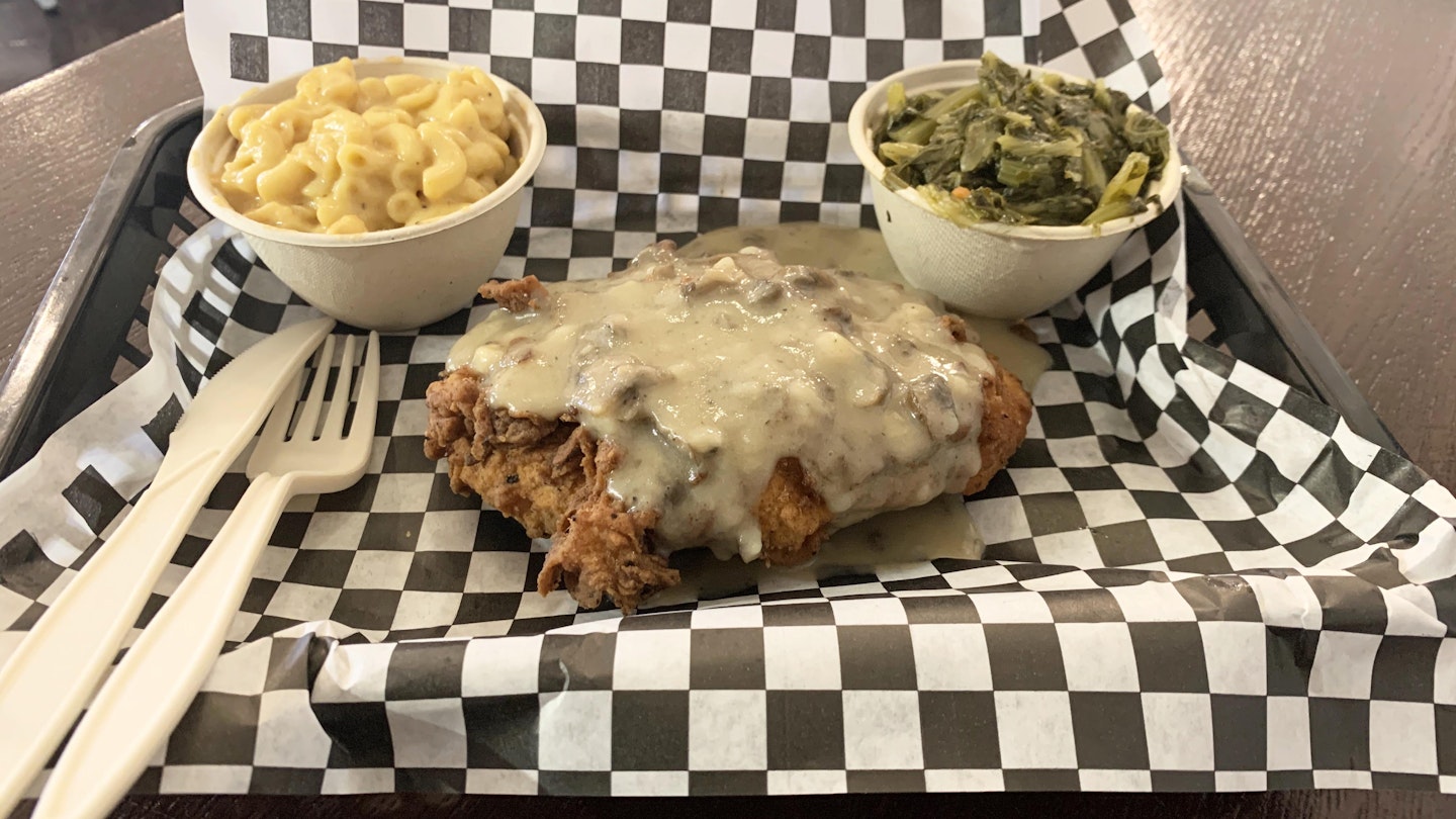 Small bowls of vegan mac and cheese and turnip greens sit on opposite sides of small plastic tray lined with black and white checkered paper. In the center is vegan fried chicken smothered in mushroom sauce. A plastic knife and fork rest on the left-hand side of the tray.