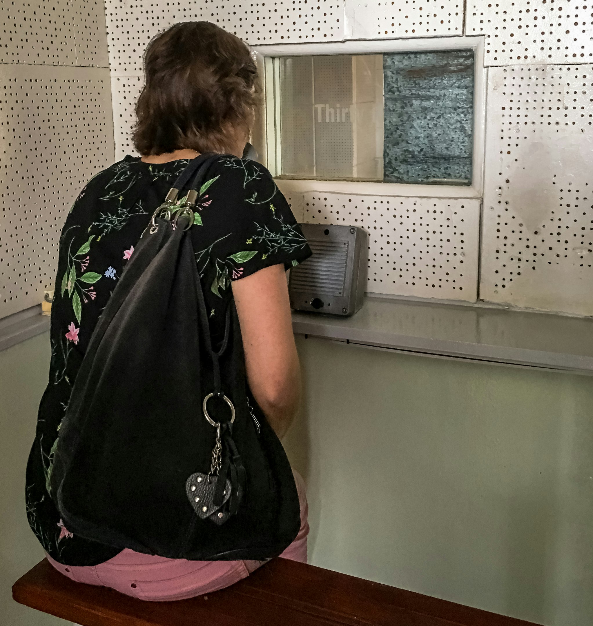 A woman sits on a wooden bench in a small room and looks through a small glass window into where a prisoner would be sitting when meeting his family.