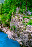 A bright blue pool within a river that is flowing between rocky cliffs topped with bright green evergreen trees.