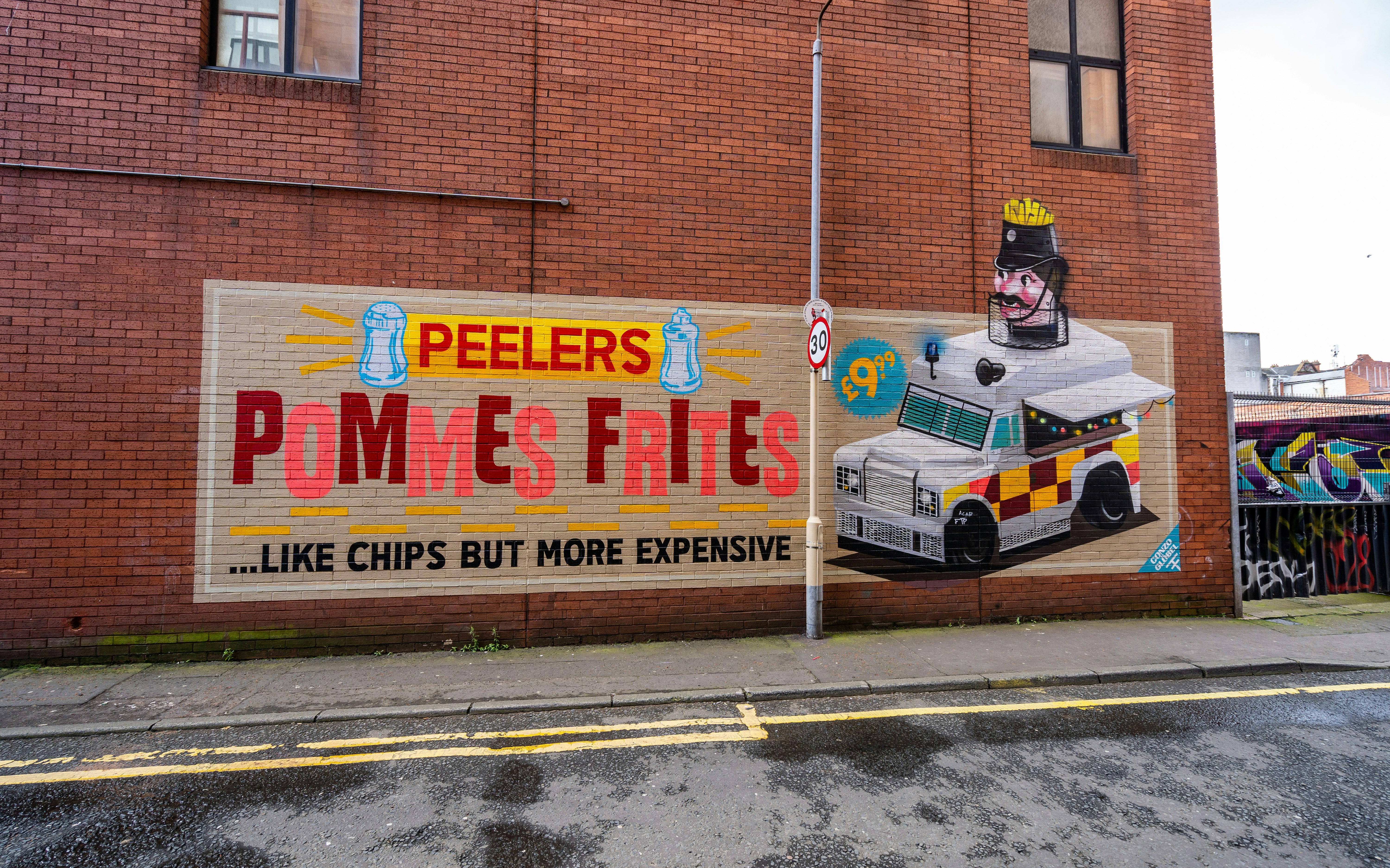 A red brick wall, painted with a colourful mural depicting an advertisement for Peeler's Pommes Frites in the style of a vintage poster. 