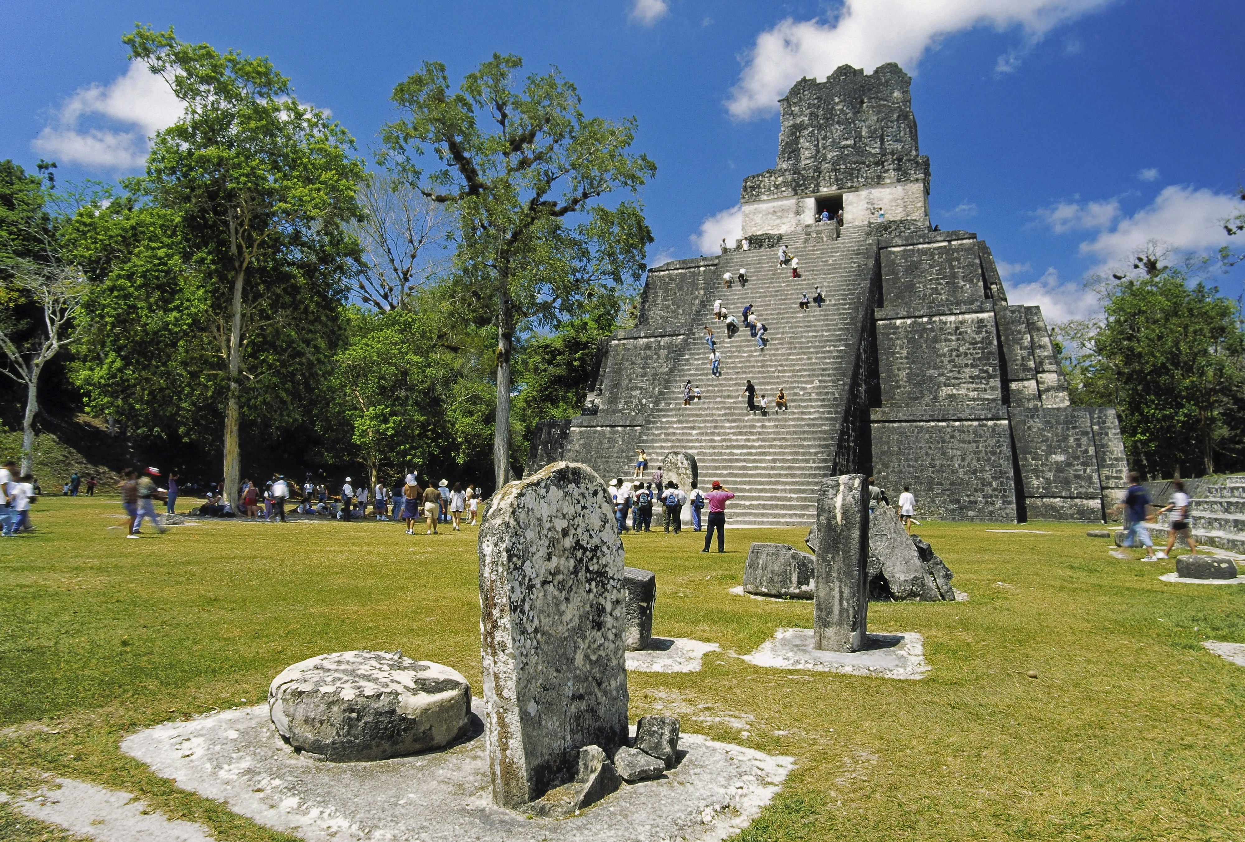 A large group of people ascend the stairs to a Mayan temple at the Tikal in Guatemala. Stone structures dot the green concourse. 