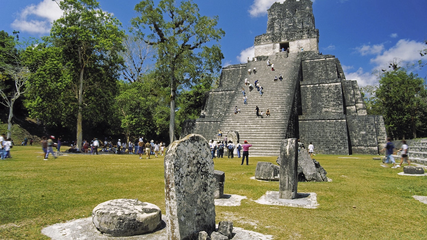A large group of people ascend the stairs to a Mayan temple at the Tikal in Guatemala. Stone structures dot the green concourse. 