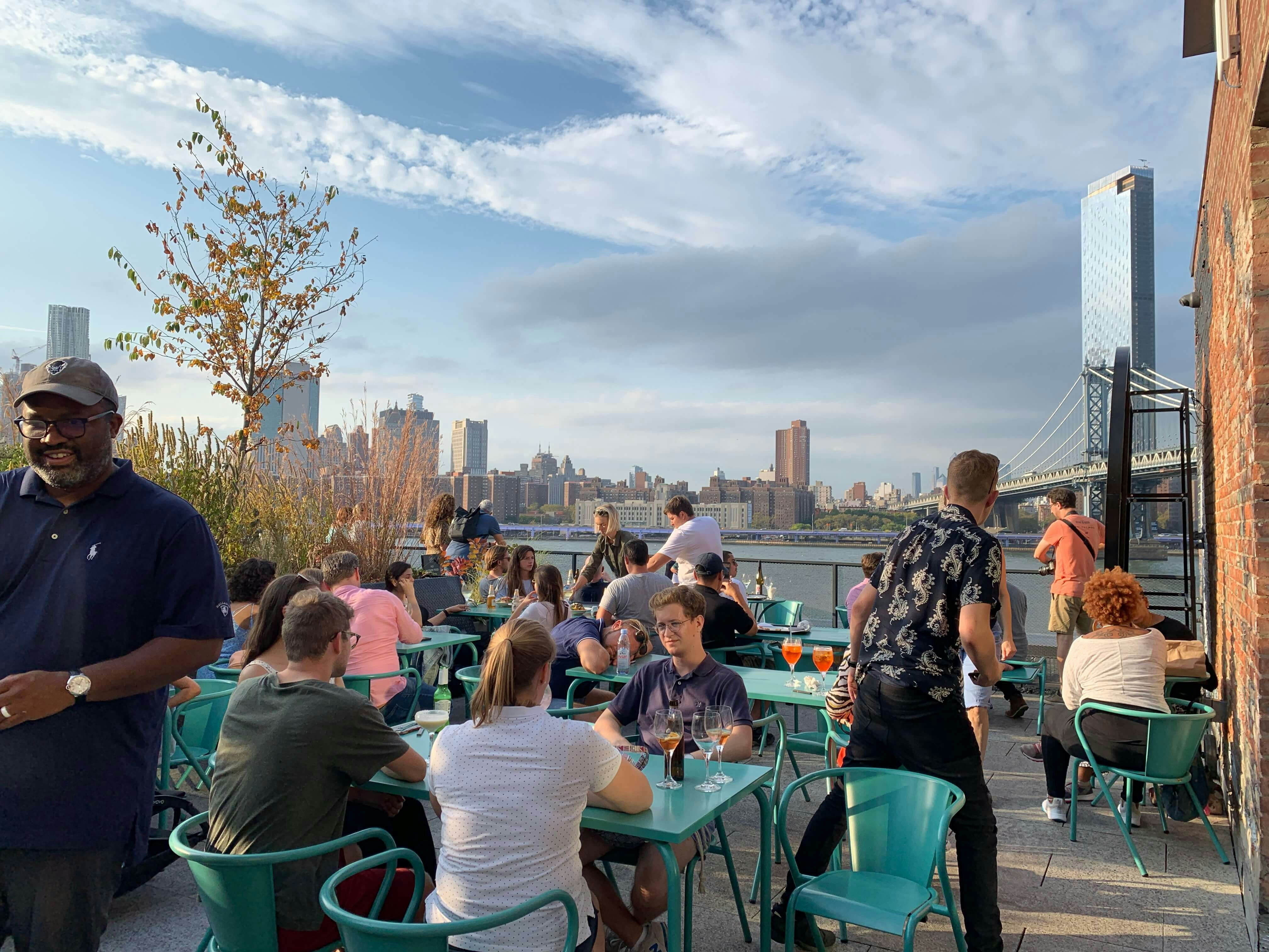 Table and chairs on an outside terrace, filled with diners. In the background is the river and views of Manhattan.