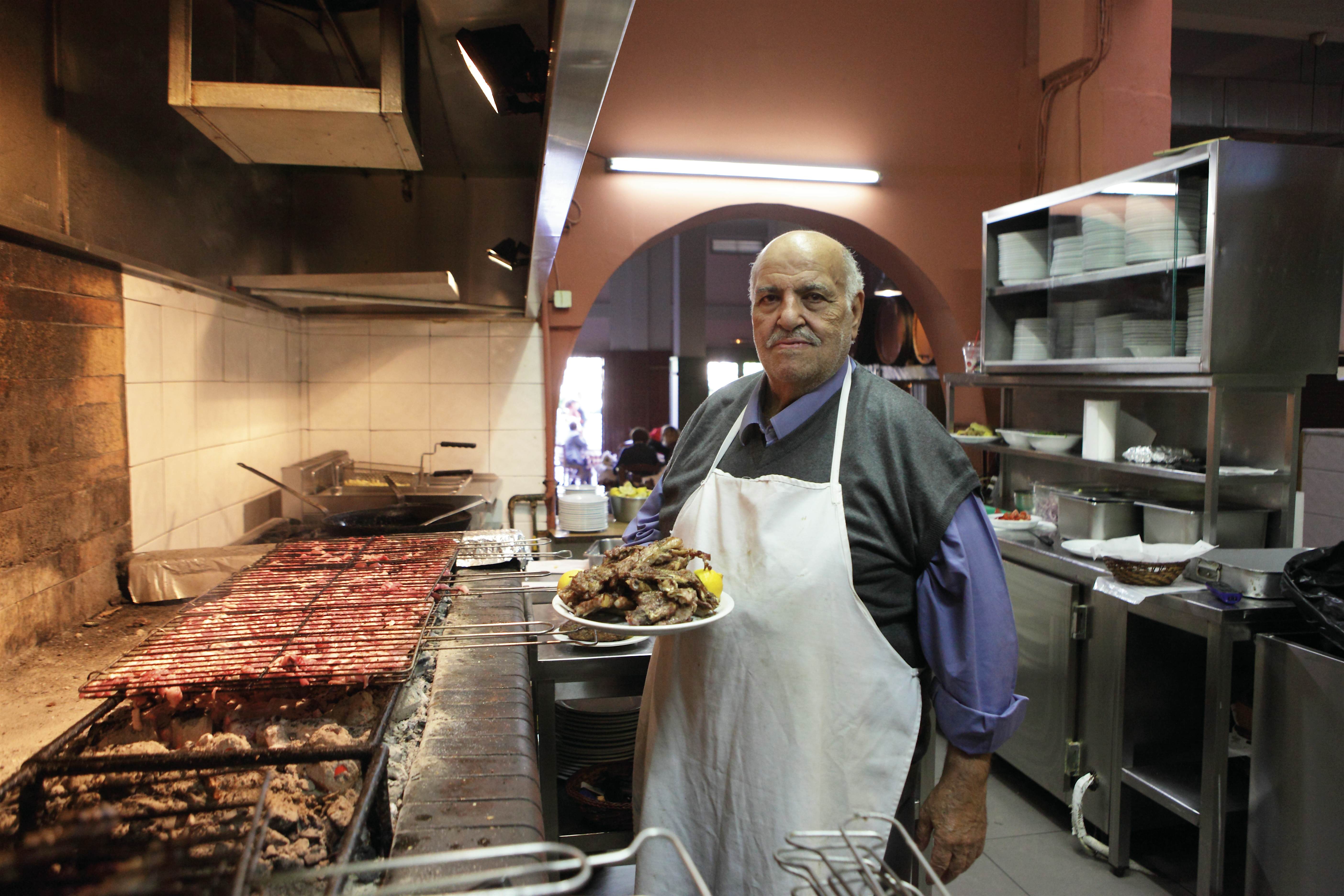 To-Steki-Tou-Elia-athens-greece-restaurants.jpg The cook at To Steki Tou Elia holds a plate of grilled meat garnished with lemon in front of massive wood-fired grills in the restaurant kitchen. He wears a blue button-down shirt covered with a sweater vest and a white apron. He looks at the camera with a proud expression.