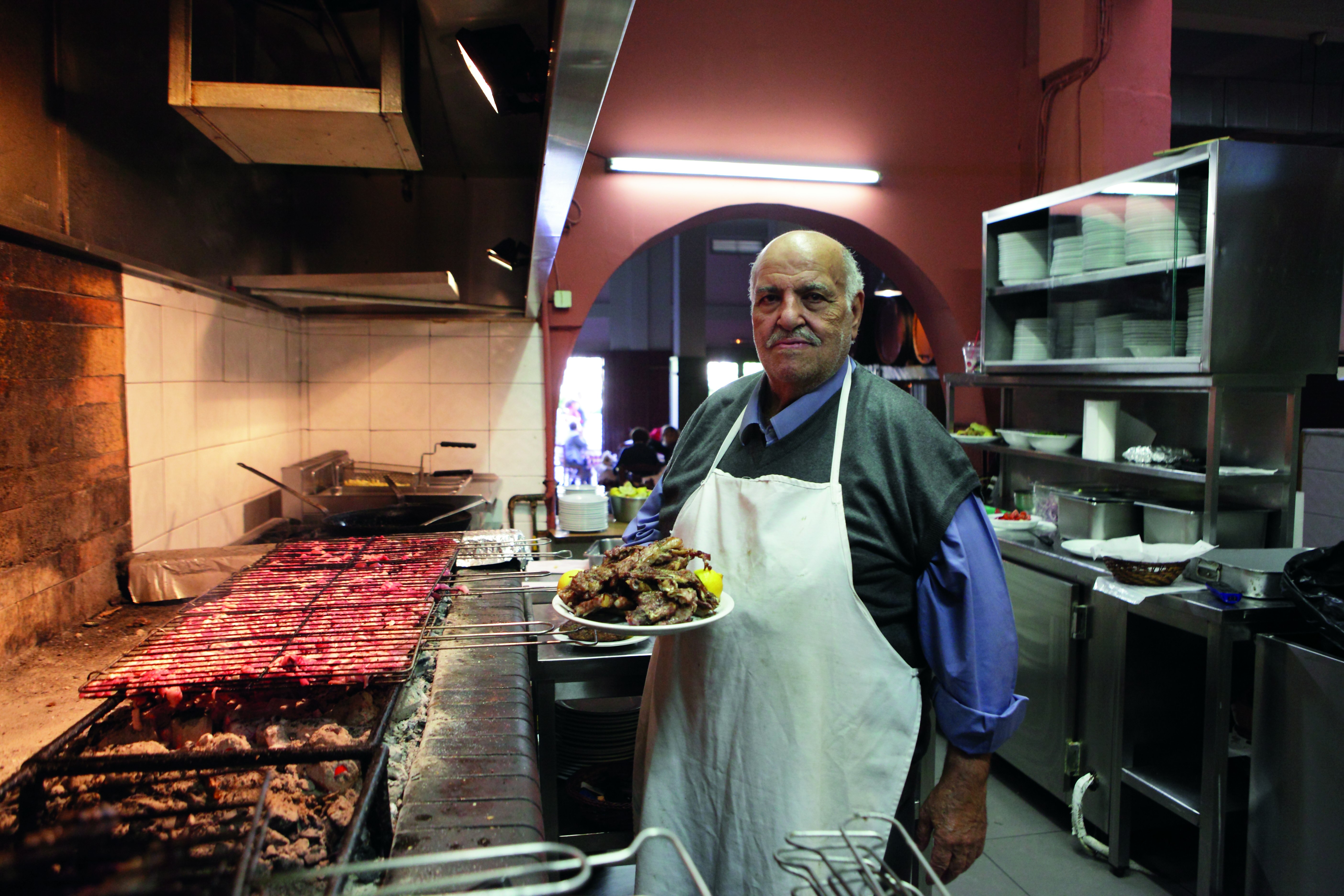 The cook at To Steki Tou Elia holds a plate of grilled meat garnished with lemon in front of massive wood-fired grills in the restaurant kitchen. He wears a blue button-down shirt covered with a sweater vest and a white apron. He looks at the camera with a proud expression.