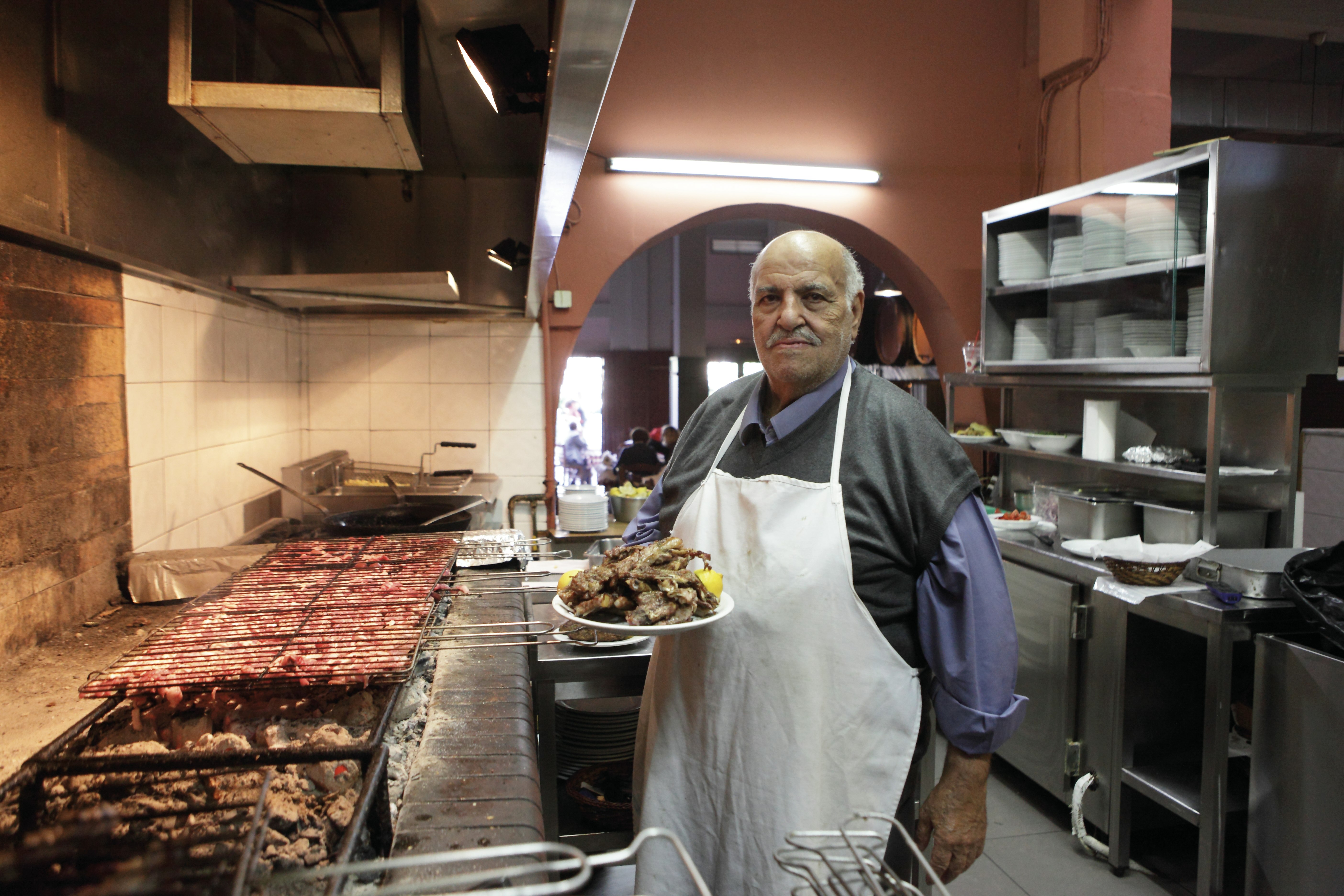 Kocken på To Steki Tou Elia håller en tallrik grillat kött garnerad med citron framför massiva vedeldade grillar i restaurangköket.  Han bär en blå button-down skjorta täckt med en tröjaväst och ett vitt förkläde.  Han tittar på kameran med ett stolt uttryck.