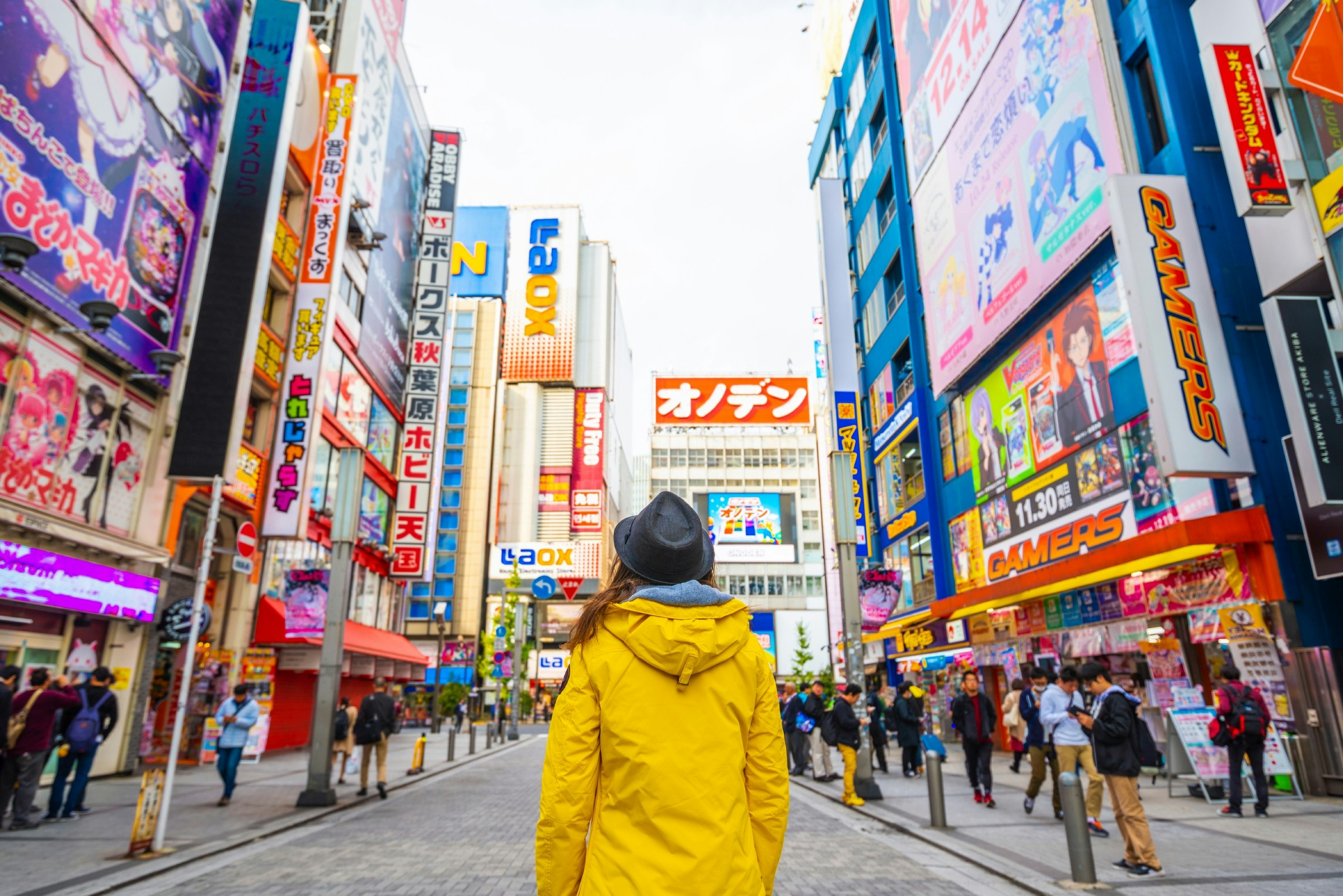 Someone wearing a bright yellow raincoat stands with their back to the camera, looking up at the colourful signs in the electronic town district of Akihabara, Tokyo. 