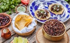 Large Mexican food spread containing corn tortillas, dried chillies, slices of lime, a small ceramic jar of green sauce, a basket full of cooke grasshoppers and two fully assembled tacos on a large blue and white plate 
