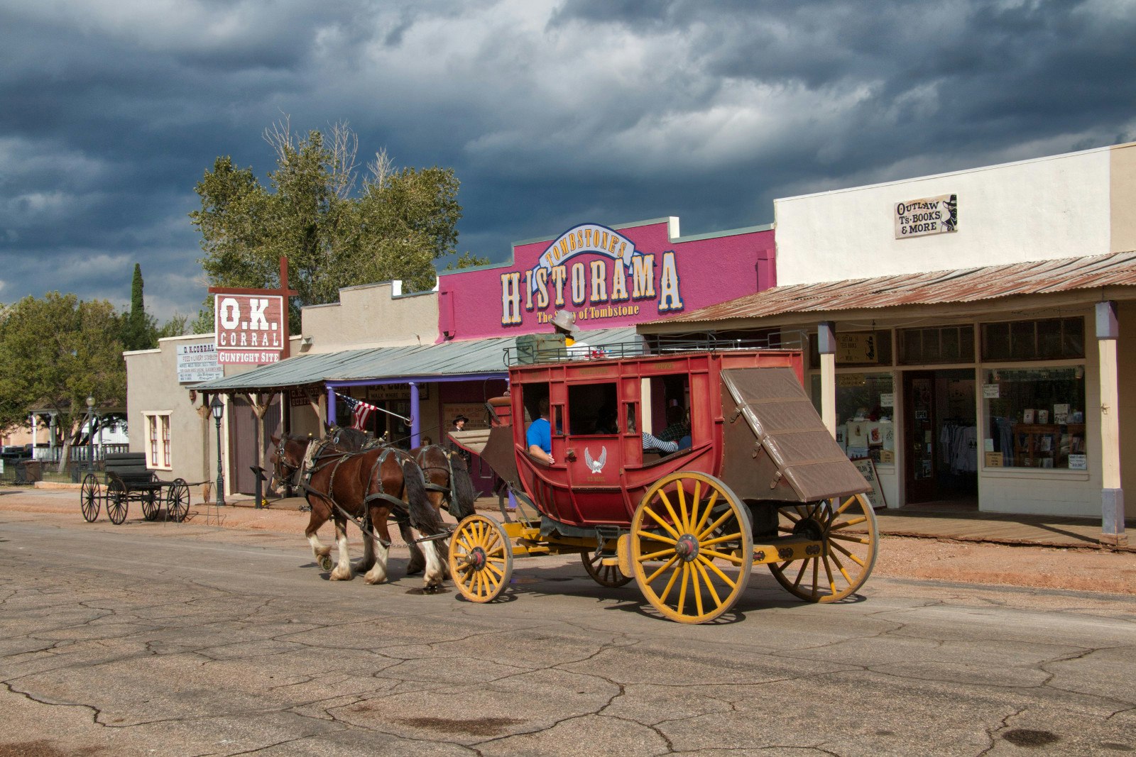 An old stage coach driven by horses goes through the town. There are storefronts behind with Wild West style signs.