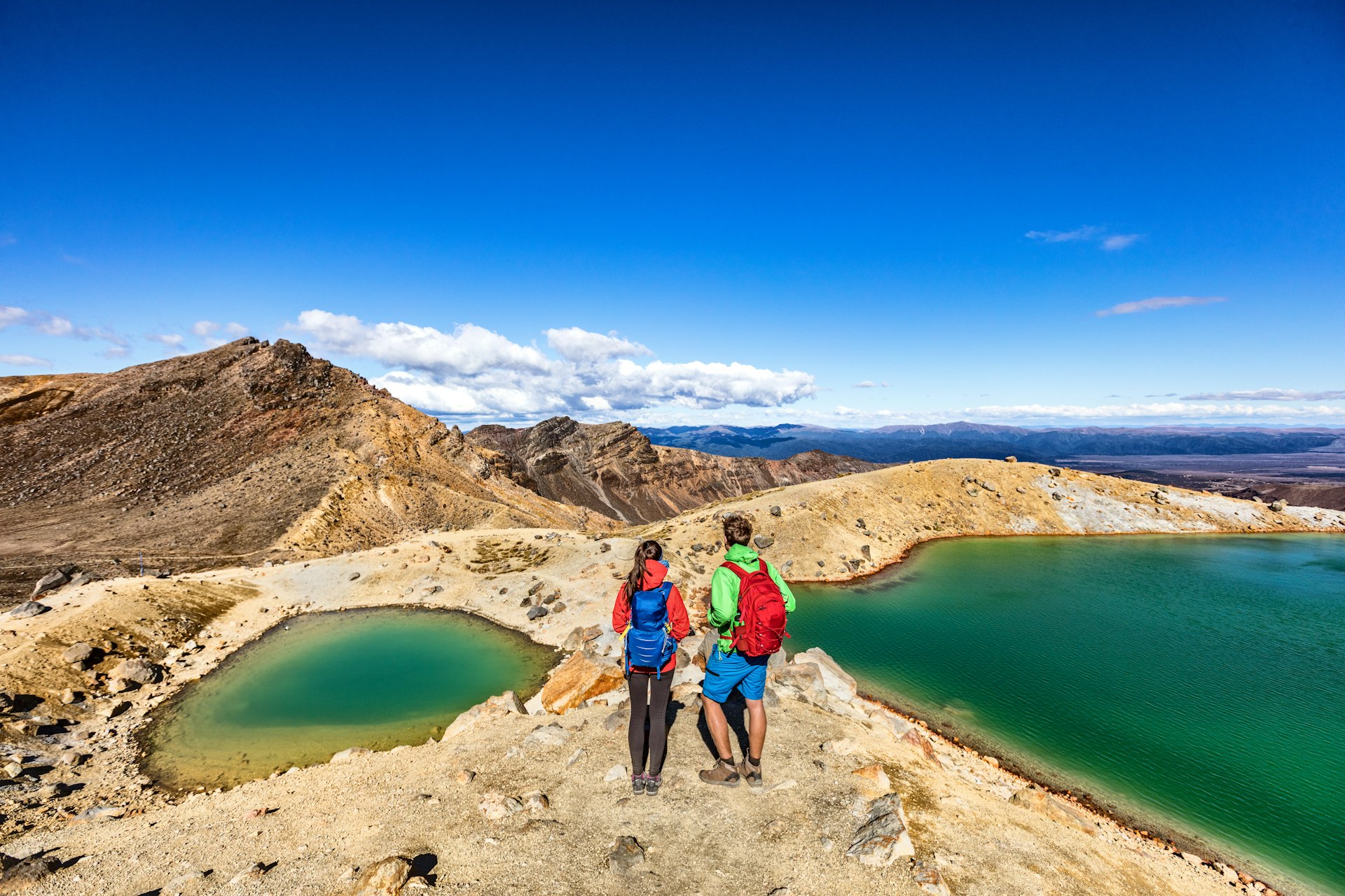 Two hikers stand between bright green lakes looking out over the landscape
