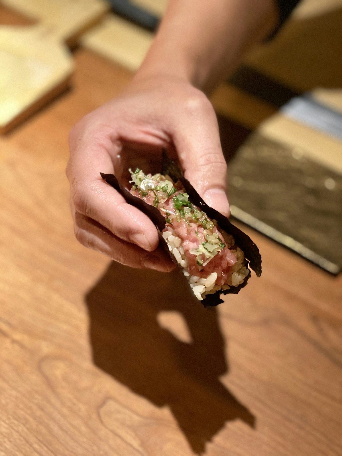 A hand holding a Toro dog, Sake no Hana. Raw tuna sits on a bed of rice, wrapped in seaweed, topped with fresh yuzu