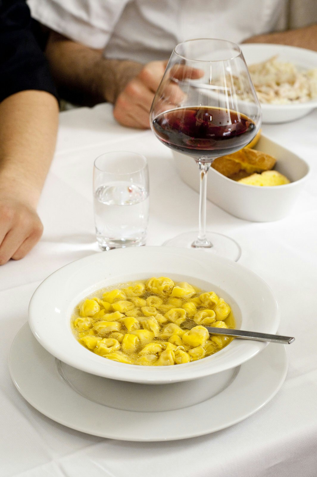 A close up of Tortellini in Brodo served in a white dish on a white tablecloth. The pasta and broth dish is next to a large glass of red wine.