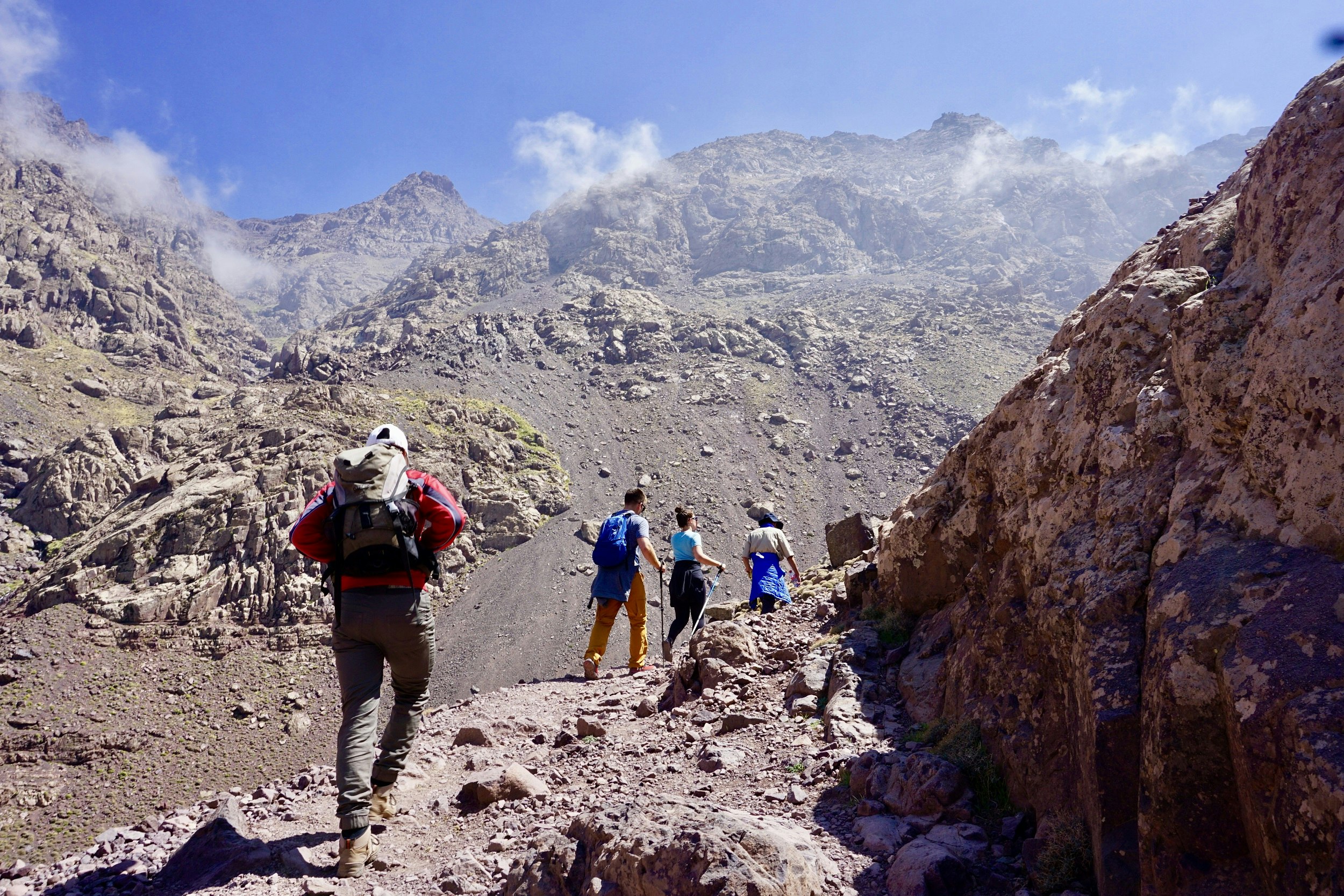Four trekkers walk up a rocky path, with the upper flanks of Toubkal rising in the background; whispy clouds float around the summit