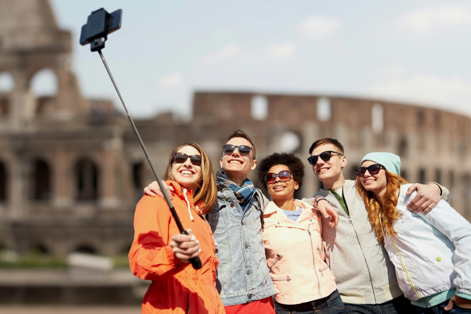 Group of smiling friends taking a selfie with smartphone and monopod with Coliseum ruins in background