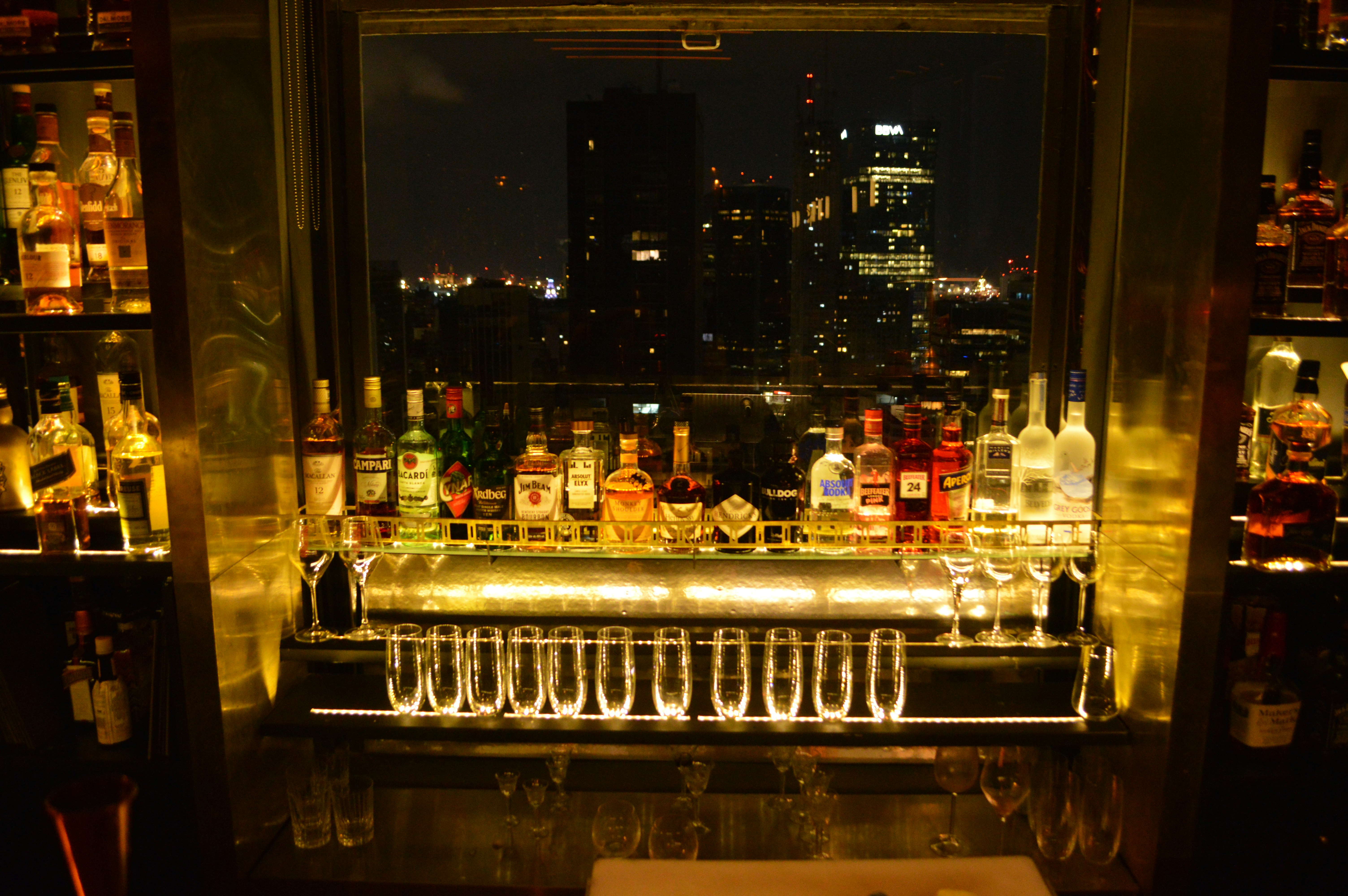 A row of liquor bottles frame a window overlooking the skyline of downtown Buenos Aires. There are a row of glasses on the lower ledge of the window that are glowing under the dim bar light