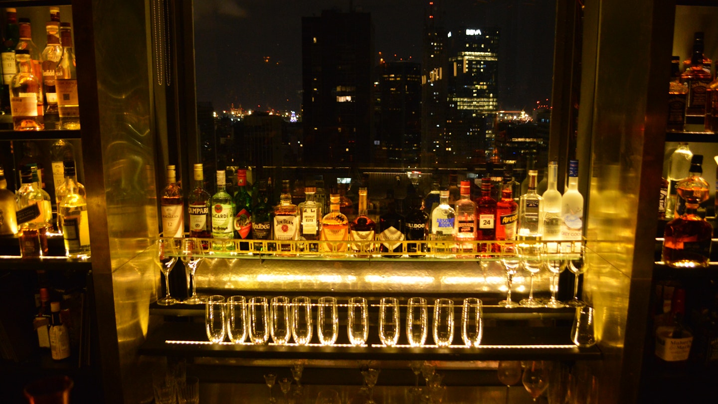 A row of liquor bottles frame a window overlooking the skyline of downtown Buenos Aires. There are a row of glasses on the lower ledge of the window that are glowing under the dim bar light