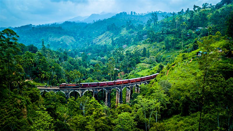 A red train crosses the Nine Arch Bridge near Ella in Sri Lanka. The stone bridge is surrounded by green, mountainous scenery on all sides. A red train crosses the Nine Arch Bridge near Ella in Sri Lanka. The stone bridge is surrounded by green, mountainous scenery on all sides.