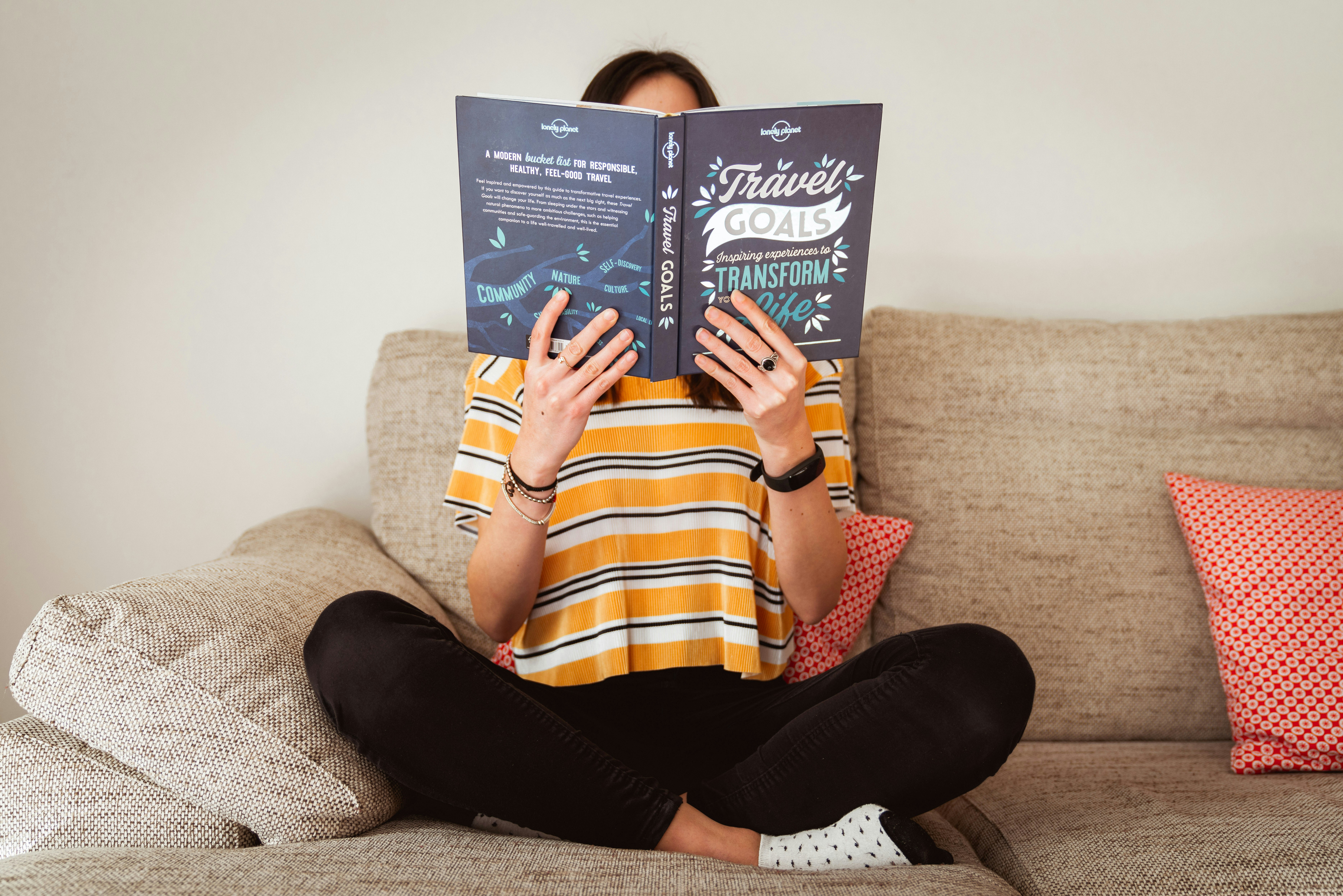 A woman is wearing black pants and a stripey yellow shirt is sitting on a couch reading Lonely Planet. Her face is obscured. 