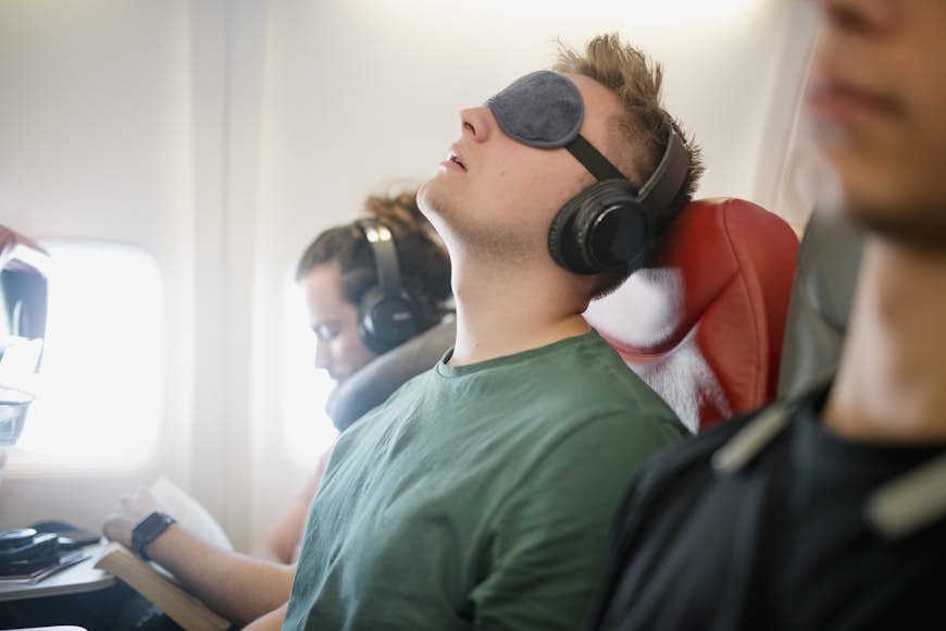 En man lutar sig bakåt i ett flygplanssäte med ögonskydd och hörlurar.  Bredvid honom står en annan man med hörlurar och nackkudde. 
