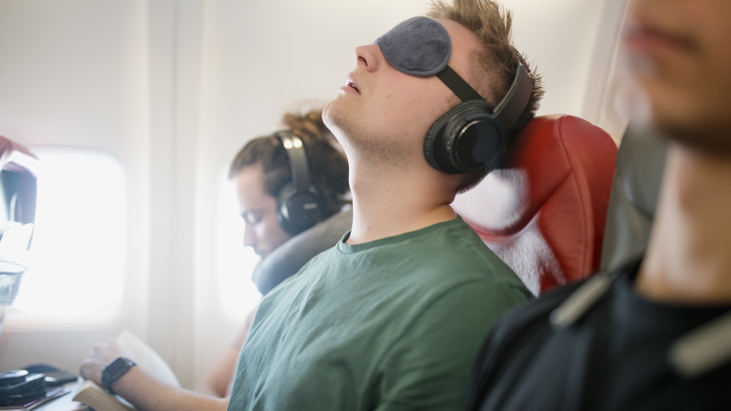 A man leans back in an airplane seat wearing an eye cover and headphones. Next to him is another man with headphones and neck pillow. 