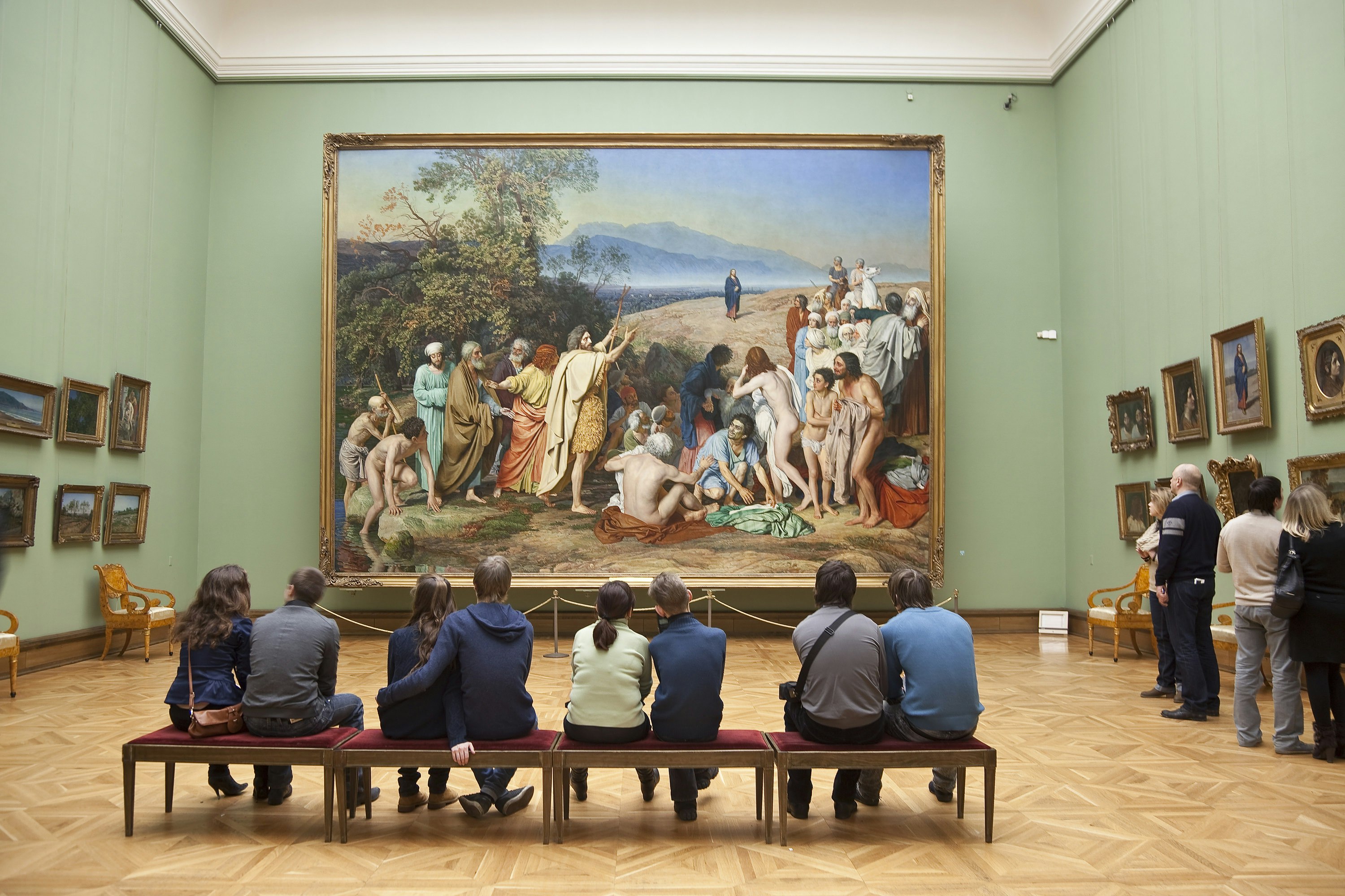 Young people sit on a bench looking at a large painting on the wall. It's by Russian artist Alexander Ivanov, called 'The appearance of Christ to the People' and is displayed in the Tretyakov gallery