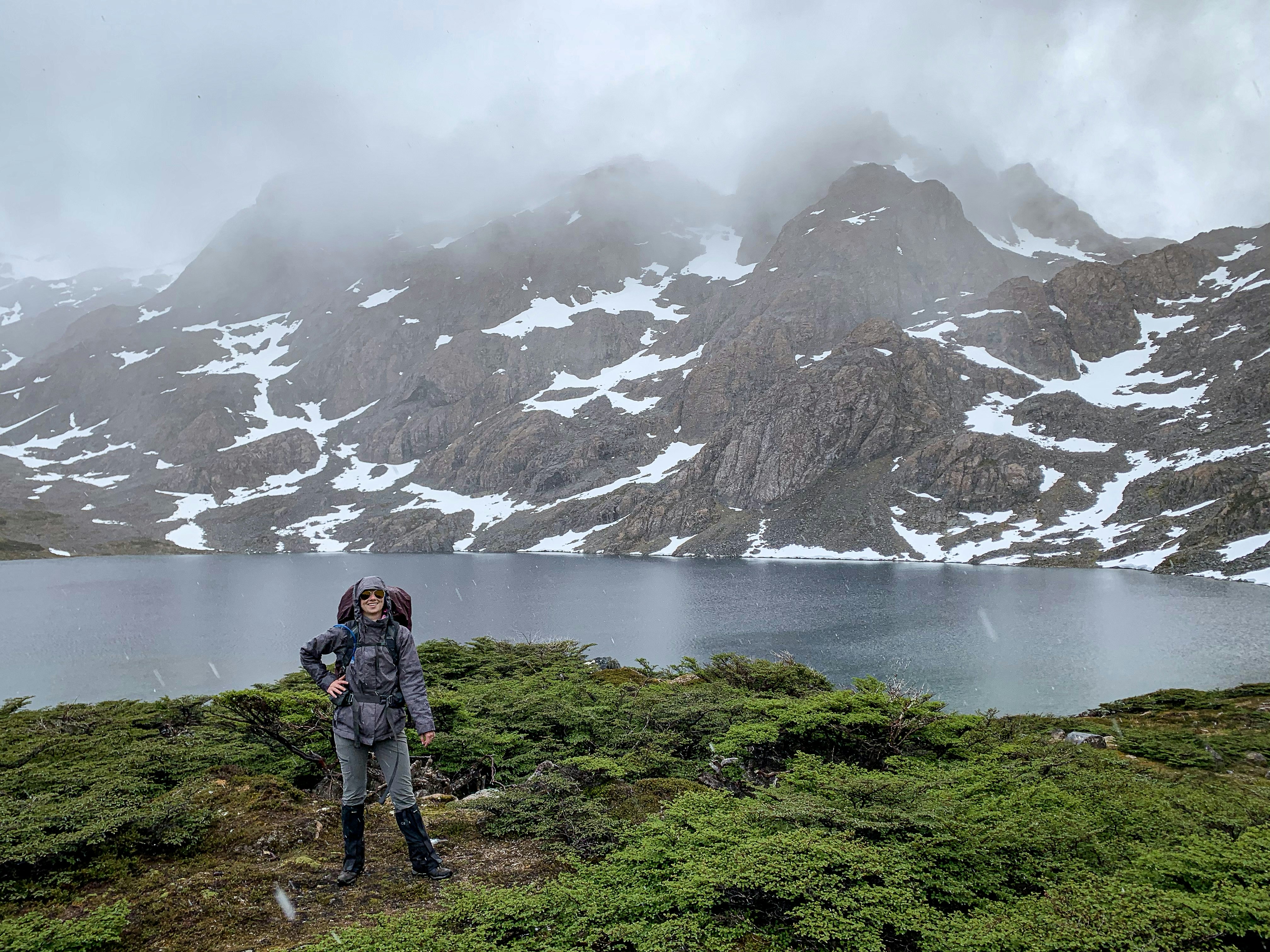 A hiker poses in front of a lake backed by snow-lined mountain slopes; sleet or rain falls