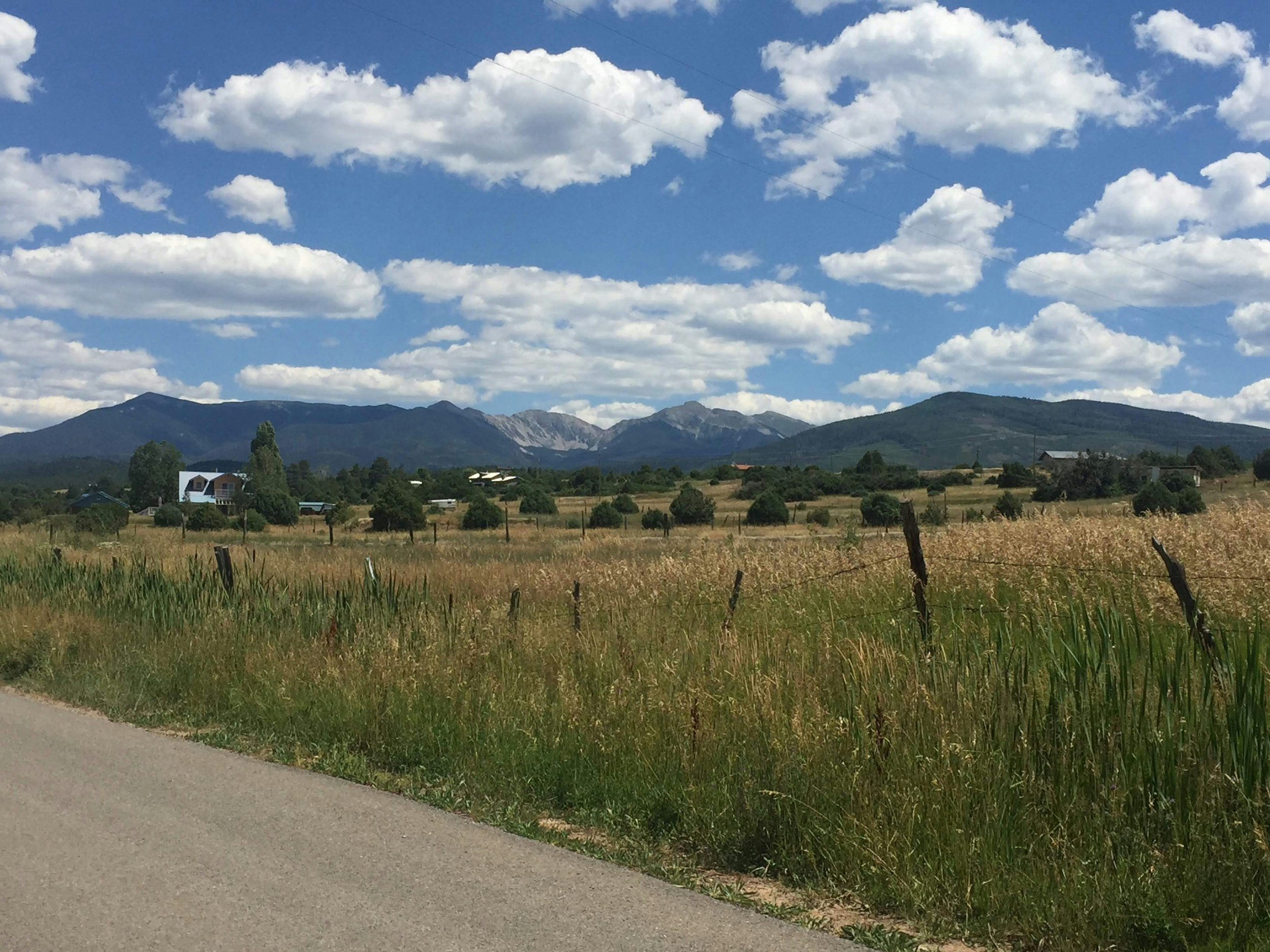 Fluffy white clouds float in a brilliant blue sky above mountains and fields on the High Road to Taos