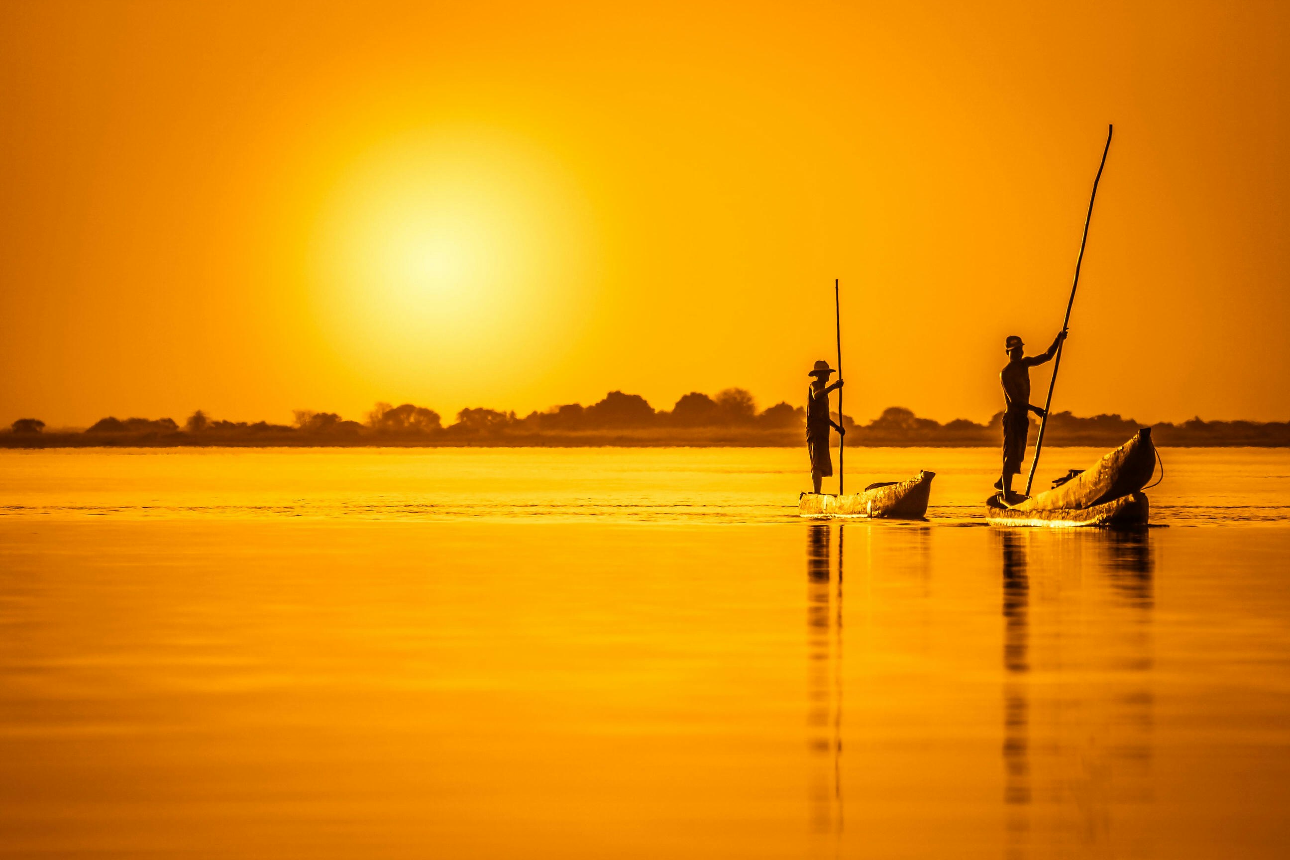 A brilliant golden image shot into the sunset, both the sky and waters of the river are the same colour; two silhouetted traditional canoes make their way on the river, both poled by standing polers.