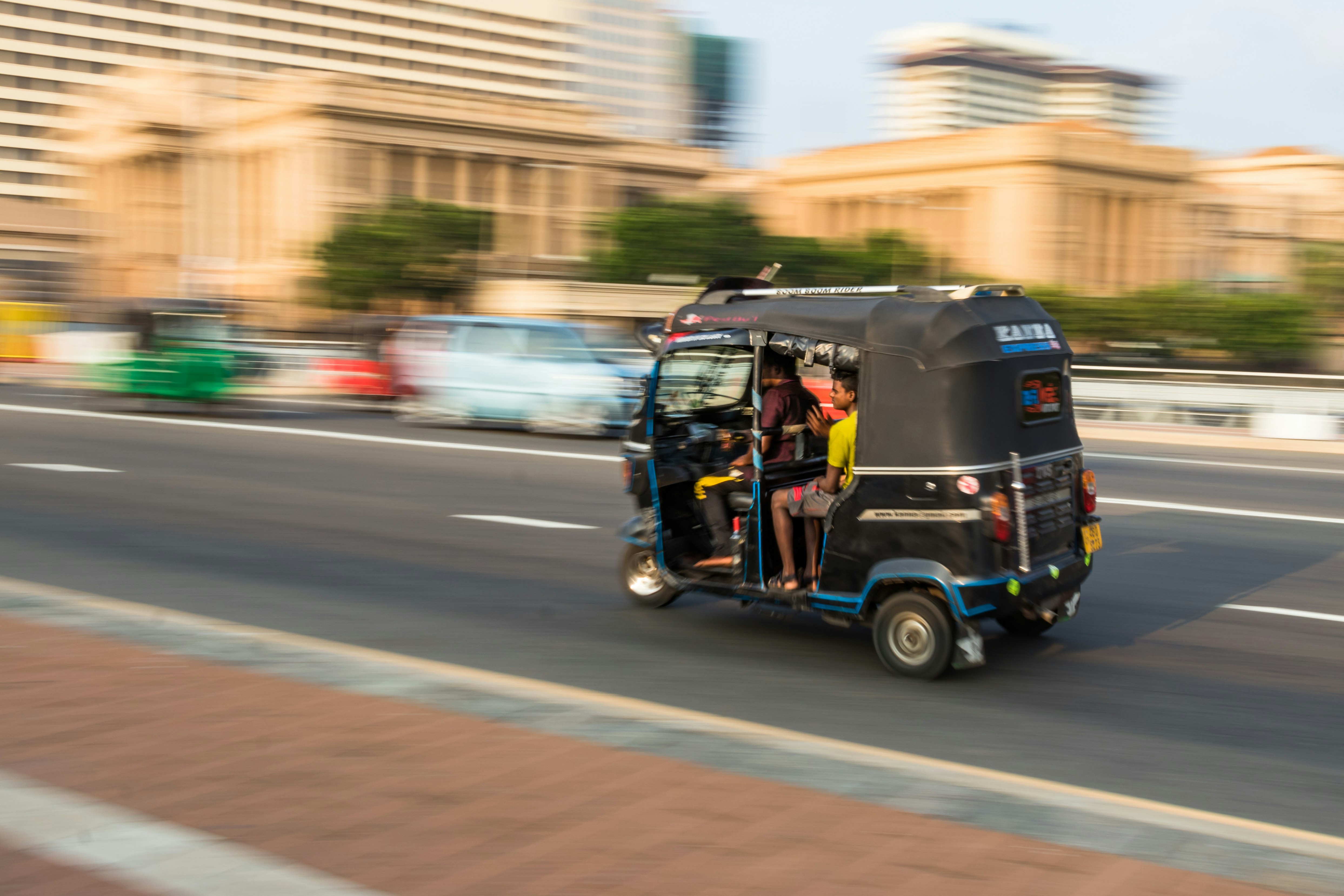 A black tuk tuk speeds along a street in Colombo, Sri Lanka. The vehicle and passengers are sharply in focus, while the background is blurred, giving the impression the tuk tuk is travelling as great speed.