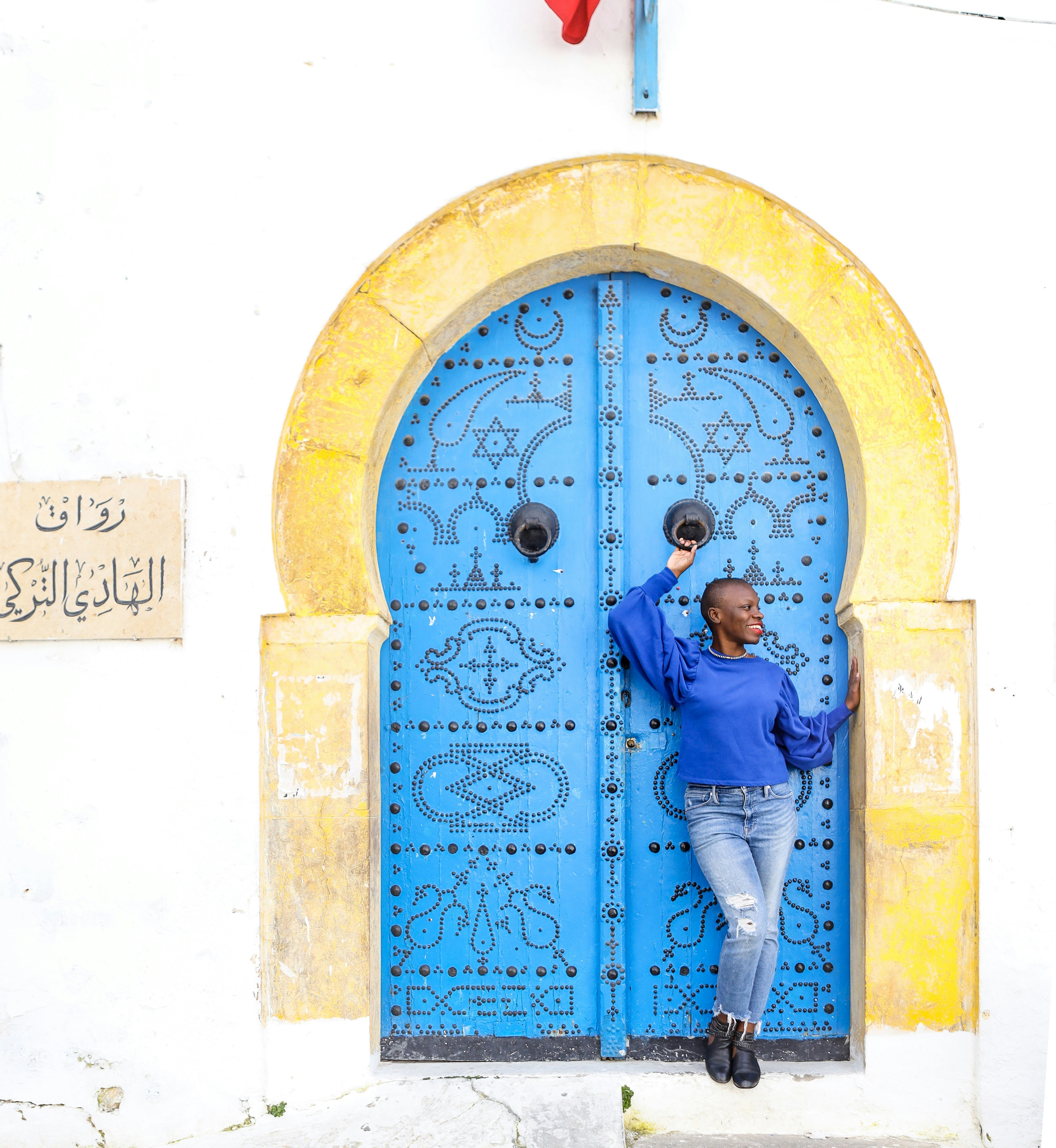 Jessica Nabongo stands in front of a large blue door with a yellow frame on he quest to travel to every country in the world