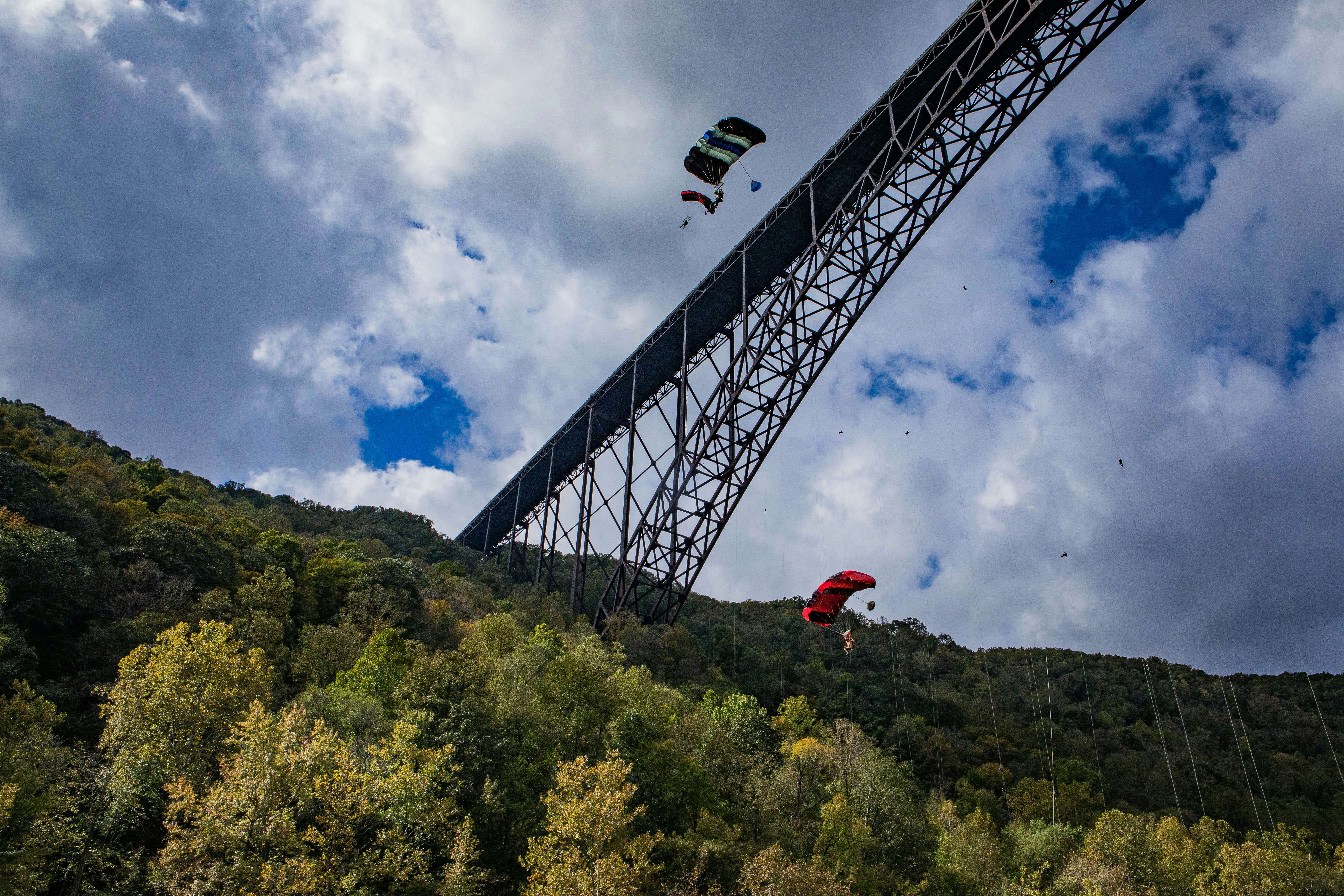 Two parachuters are descending down the side of New River Gorge Bridge. The camera is almost directly beneath them. A cloudy sky and woodland is visible.