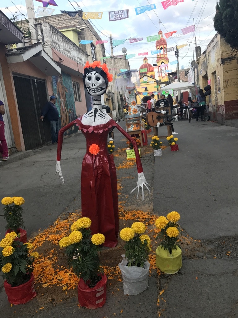 A paper mache figure of a skeletal woman wearing a Victorian-era red dress with an orange flower at the belt stands in the middle of a sidewalk. Behind her are other paper mache figures celebrating Dia de Muertos.   