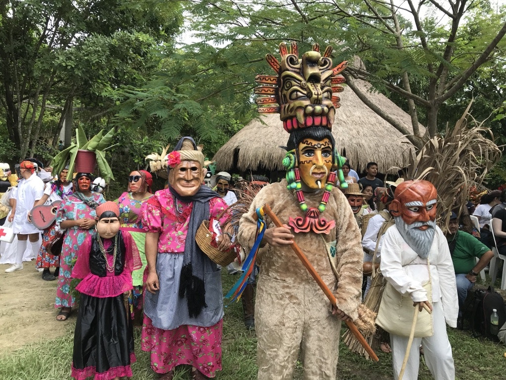 A procession of people wearing elaborate masks and traditional brightly colored clothing makes it way through a village; Dia de Muertos 