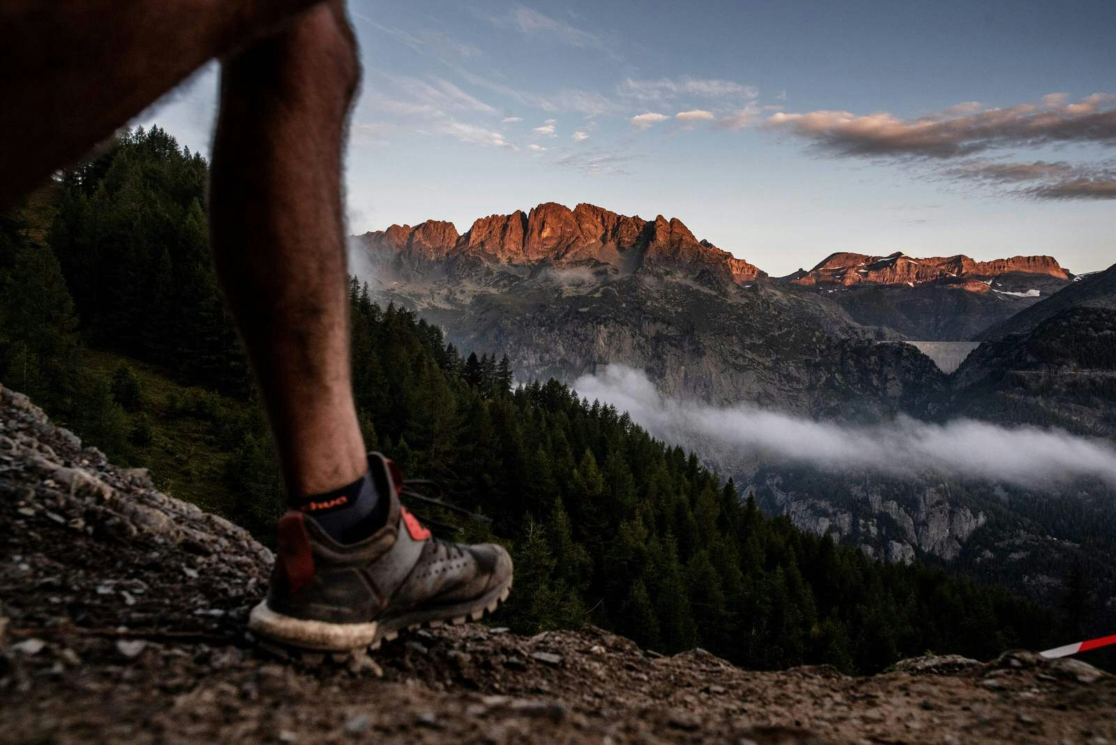 An incredible mountain ridge lit by the morning sun is the backdrop to the lower half of a runner's leg striding through the grass in the foreground