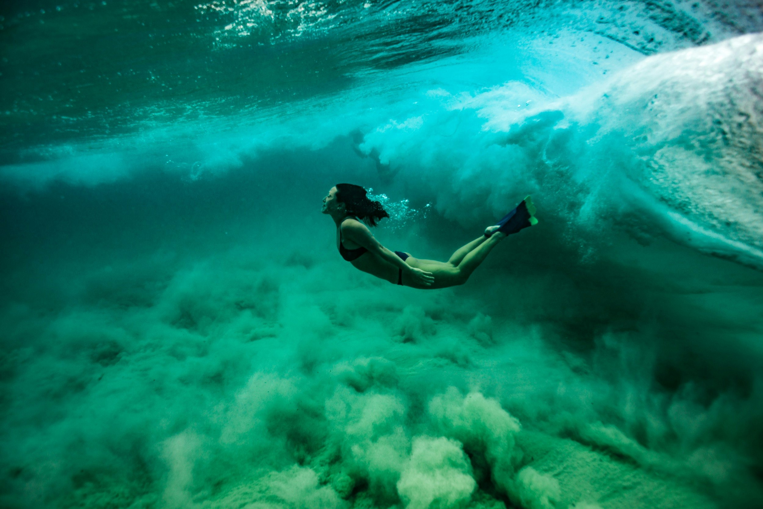 A woman floats underwater with her back arched as a wave crashes behind her head