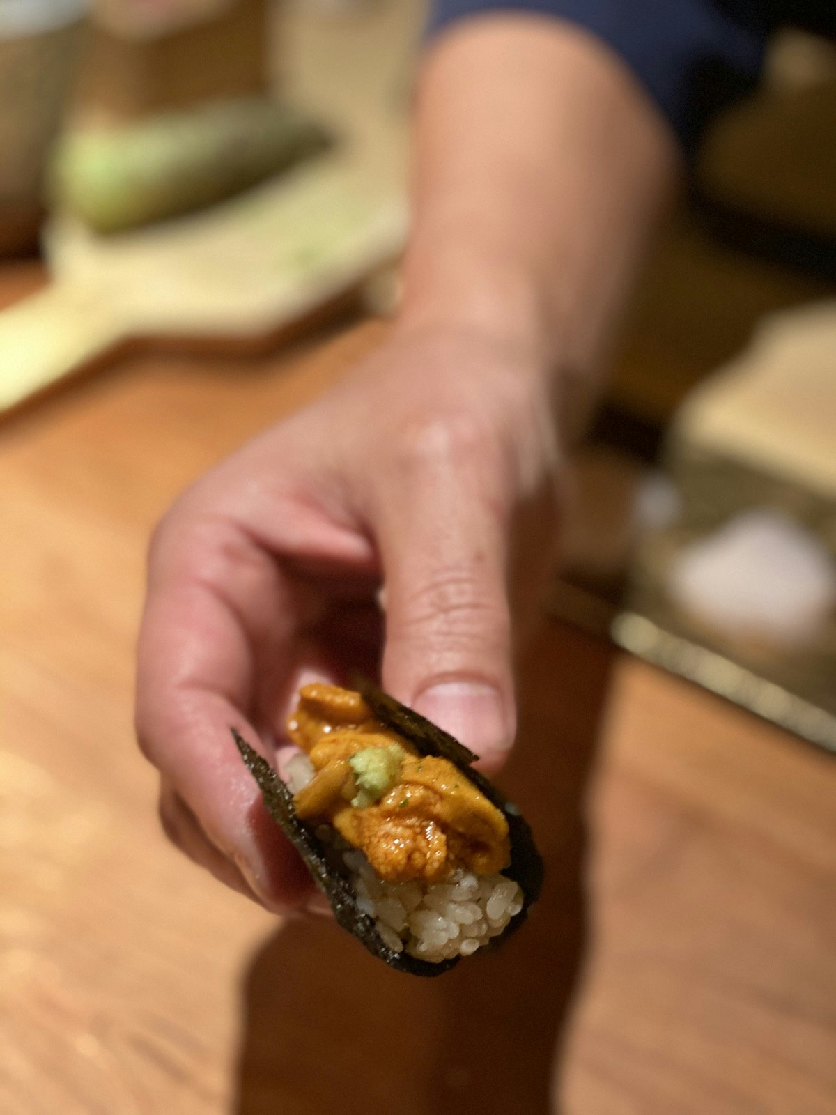 A hand holding Uni Gunkan at Sake no Hana. Orange sea urchin sits on a bed of rice wrapped in seaweed topped with fresh wasabi