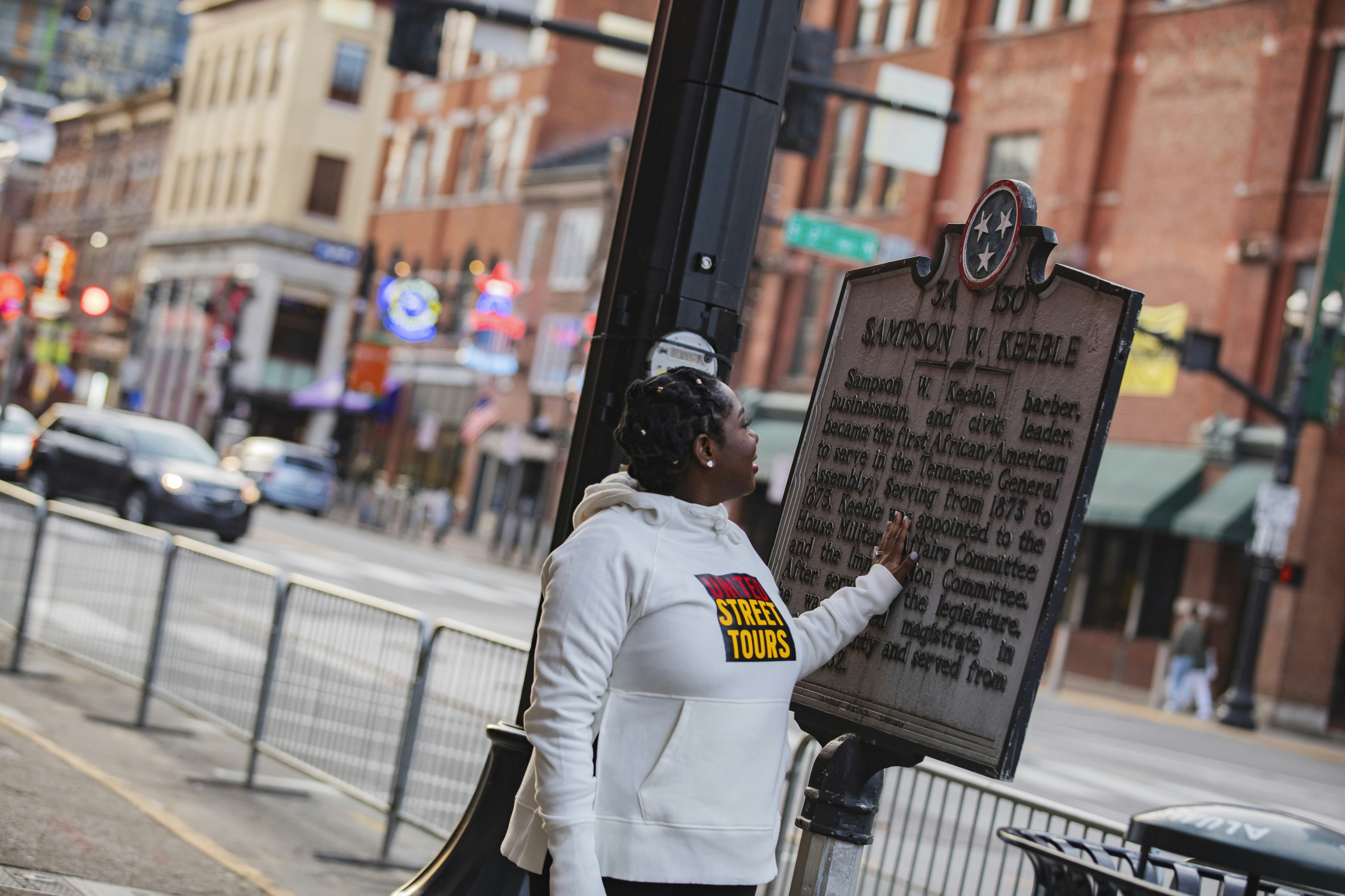 A woman wearing a white sweatshirt that says 'United Street Tours' touches a sign memorializing Sampson W. Keeble on a busy sidewalk in downtown Nashville.
