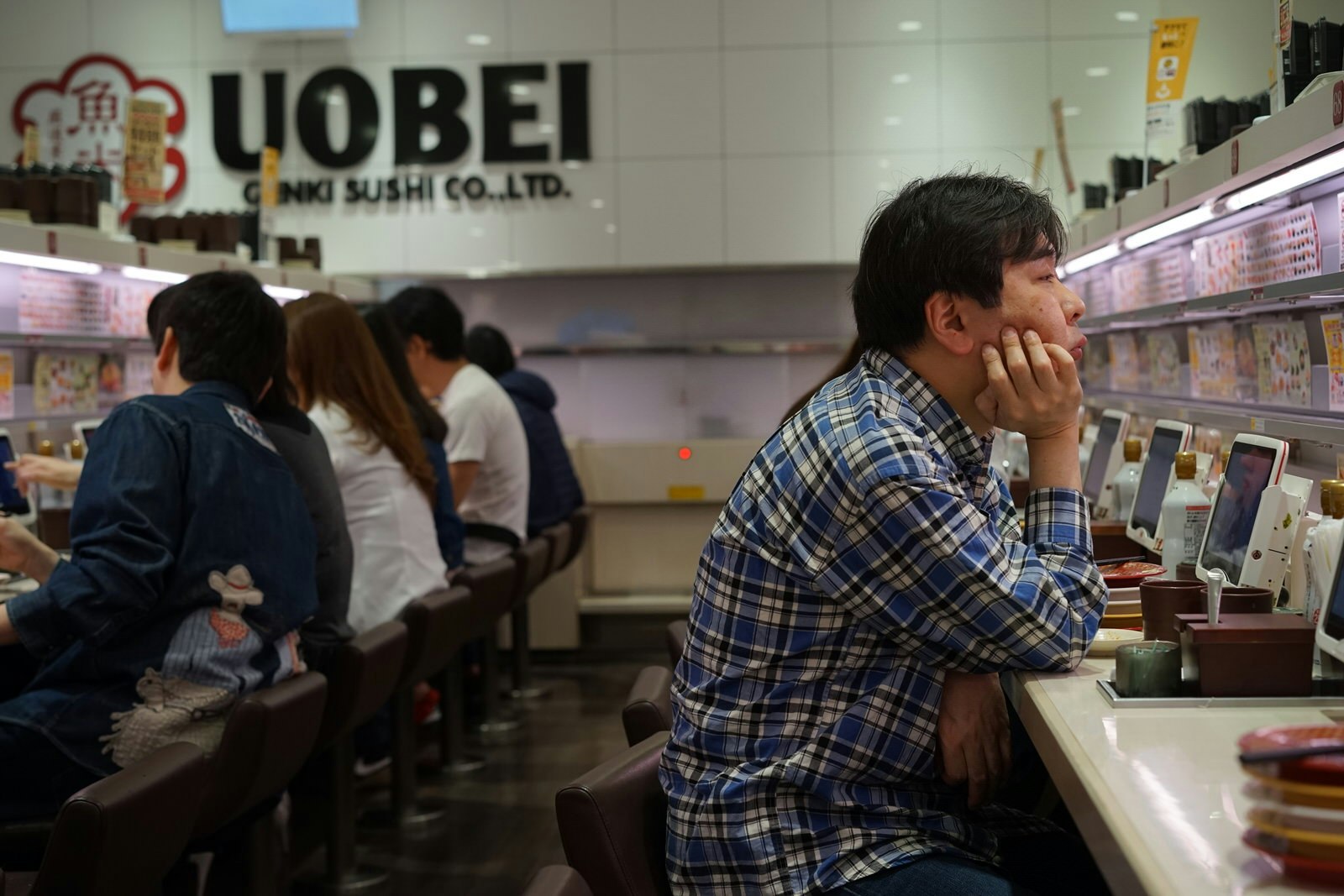 The interior of sushi restaurant Uobei. The white tiled walls feature iPads for customers to order their food on. The walls are lined with stools, filled with customers eating and chatting with each other. 