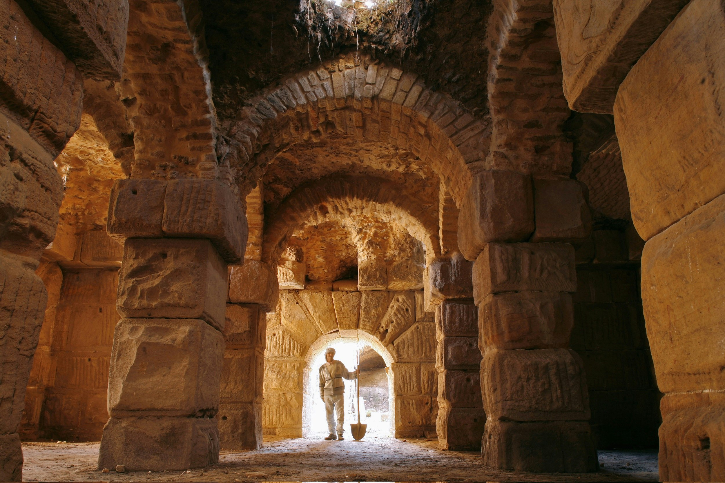 A Tunisian man stands holding a shovel at the end of a subterranean catacomb; the inside is constructed of huge, but roughly-hewn stones and brick arches. 
