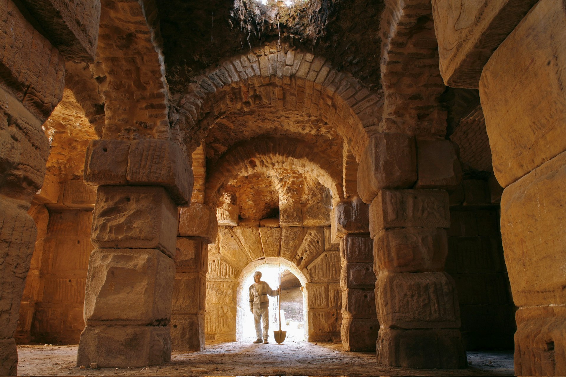 A Tunisian man stands holding a shovel at the end of a subterranean catacomb; the inside is constructed of huge, but roughly-hewn stones and brick arches. 