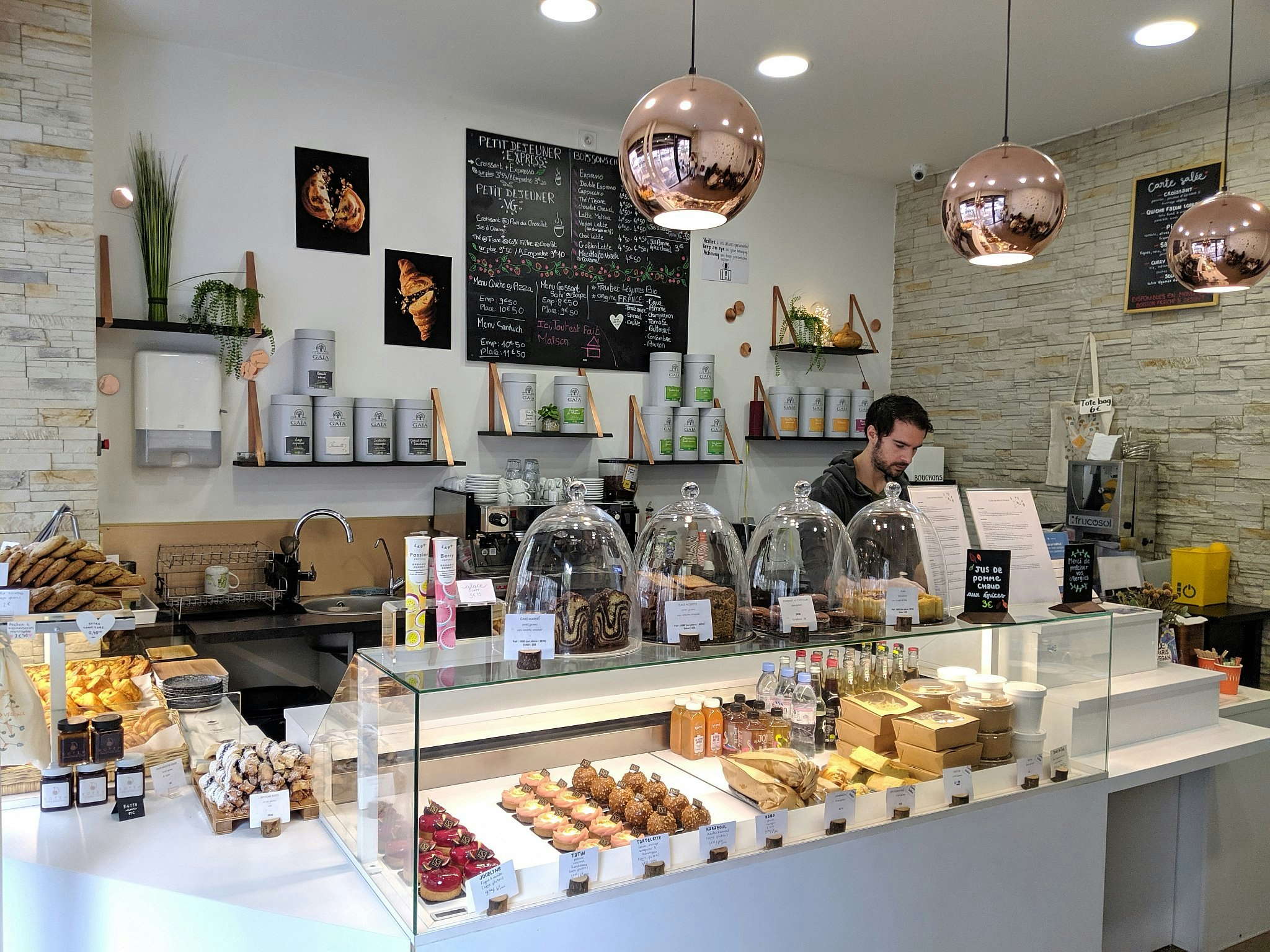 A worker behind a counter laden with cakes and sweet treats; walls are white or exposed brick with metal lights hanging from above. 