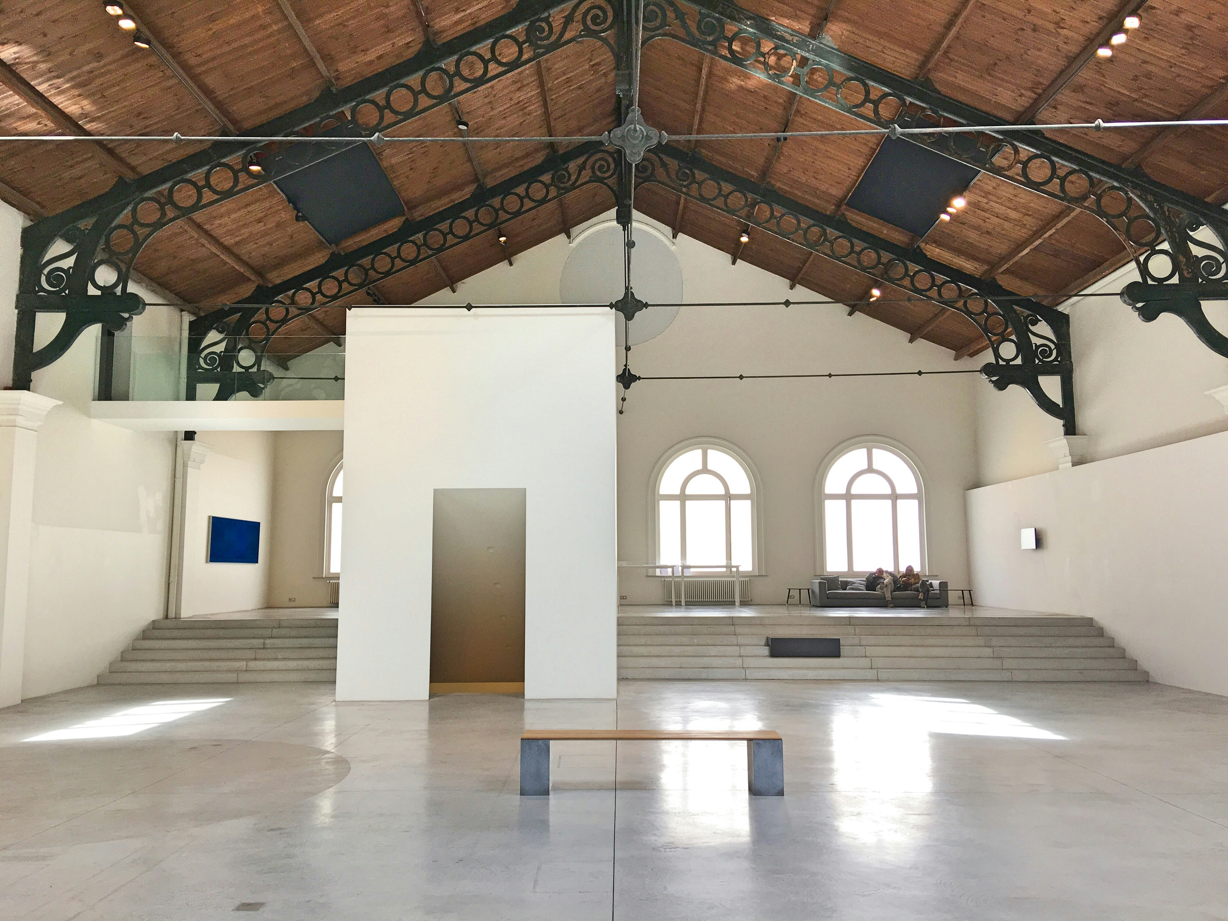 A large white room under a sloping wooden roof supported by handsome iron girders; in the middle of the room is a bench, with what looks like a freestanding doorway behind it; at the rear of the room, steps lead up to arched windows.
