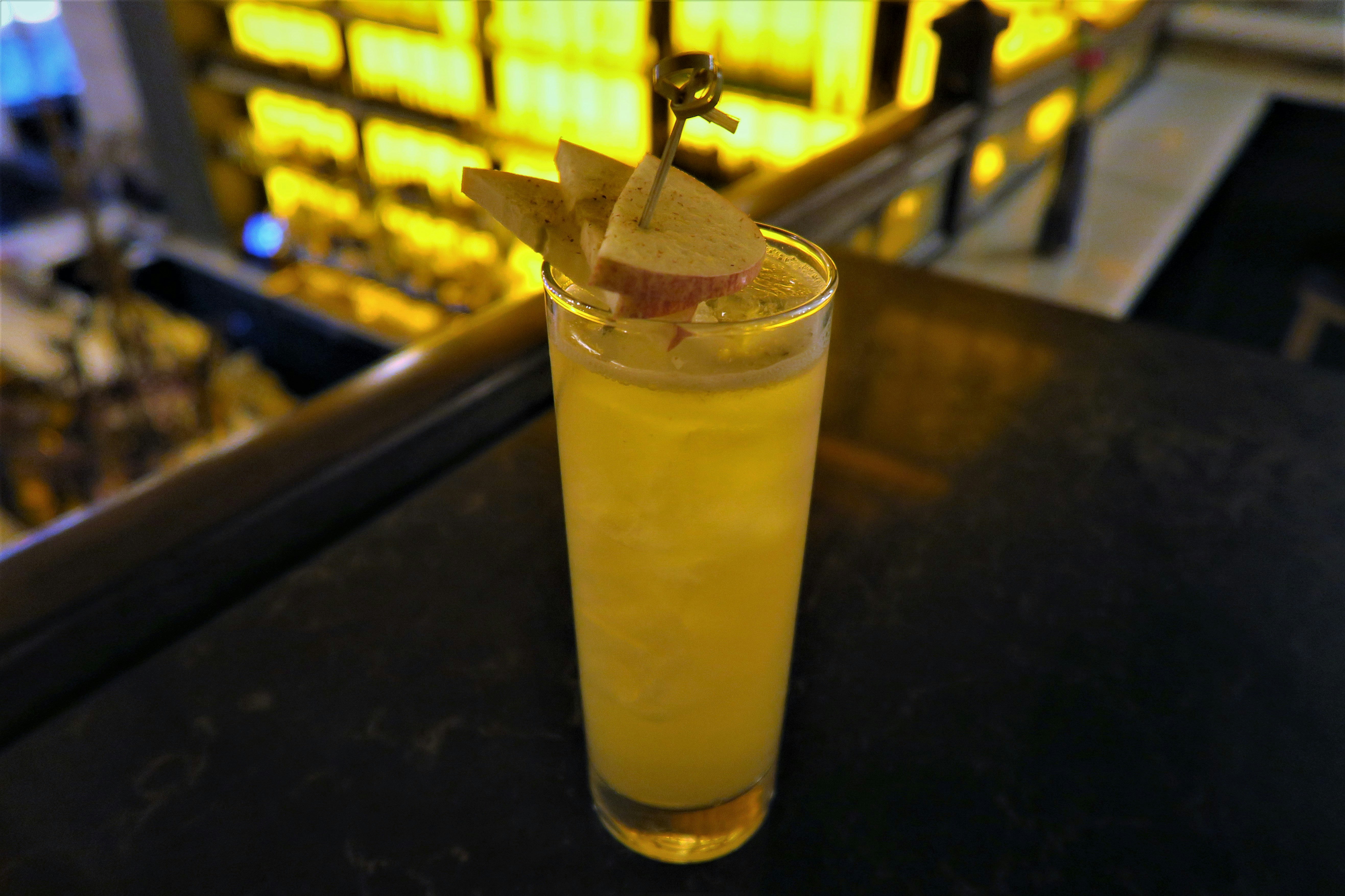 A yellow mocktail in a tall glass has a wooden stirrer and slices of fresh apple.