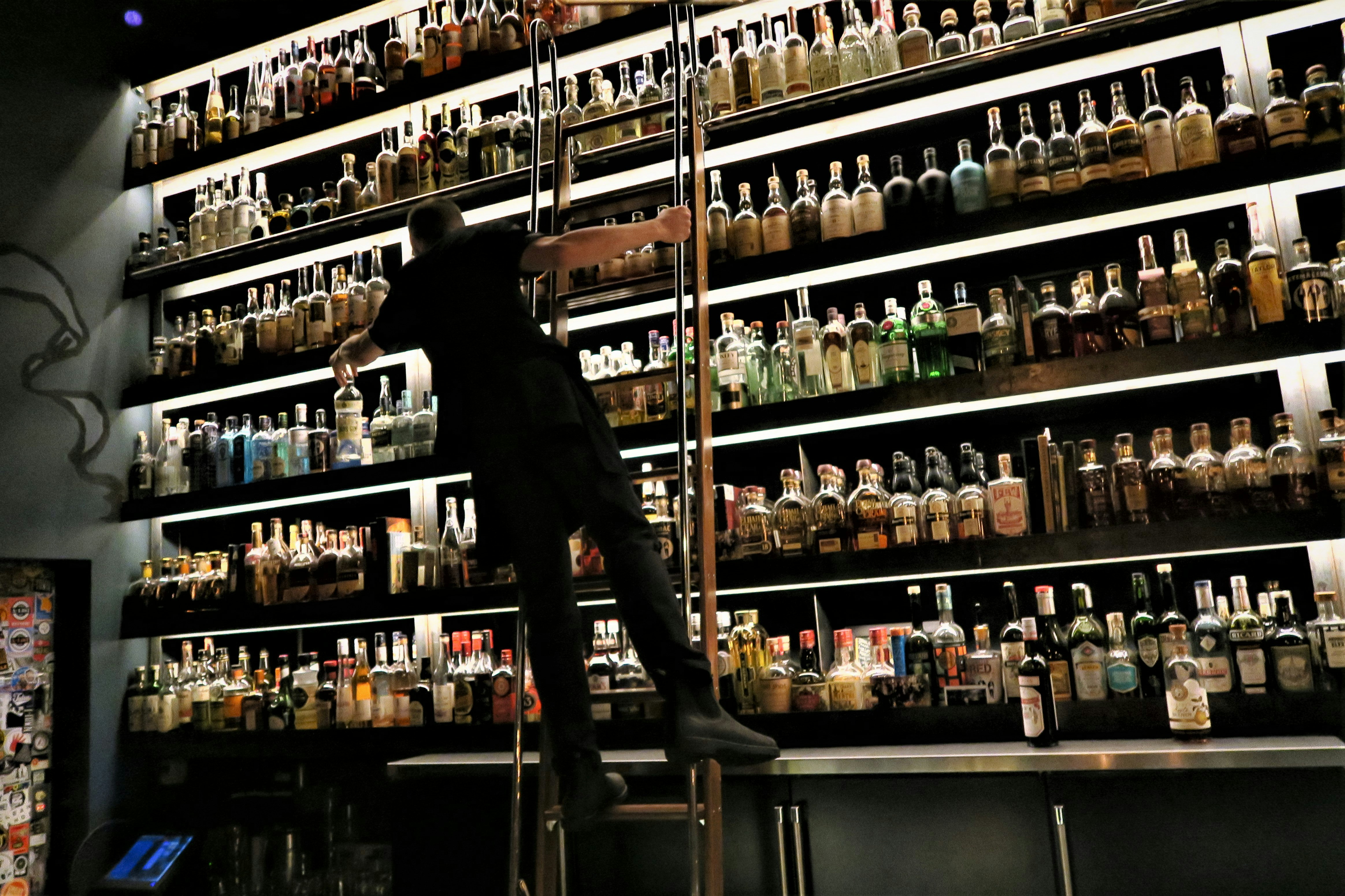 A bartender dressed in black holds on to the side of a ladder while reaching for a bottle of spirits from a multi-level shelf filled with bottles