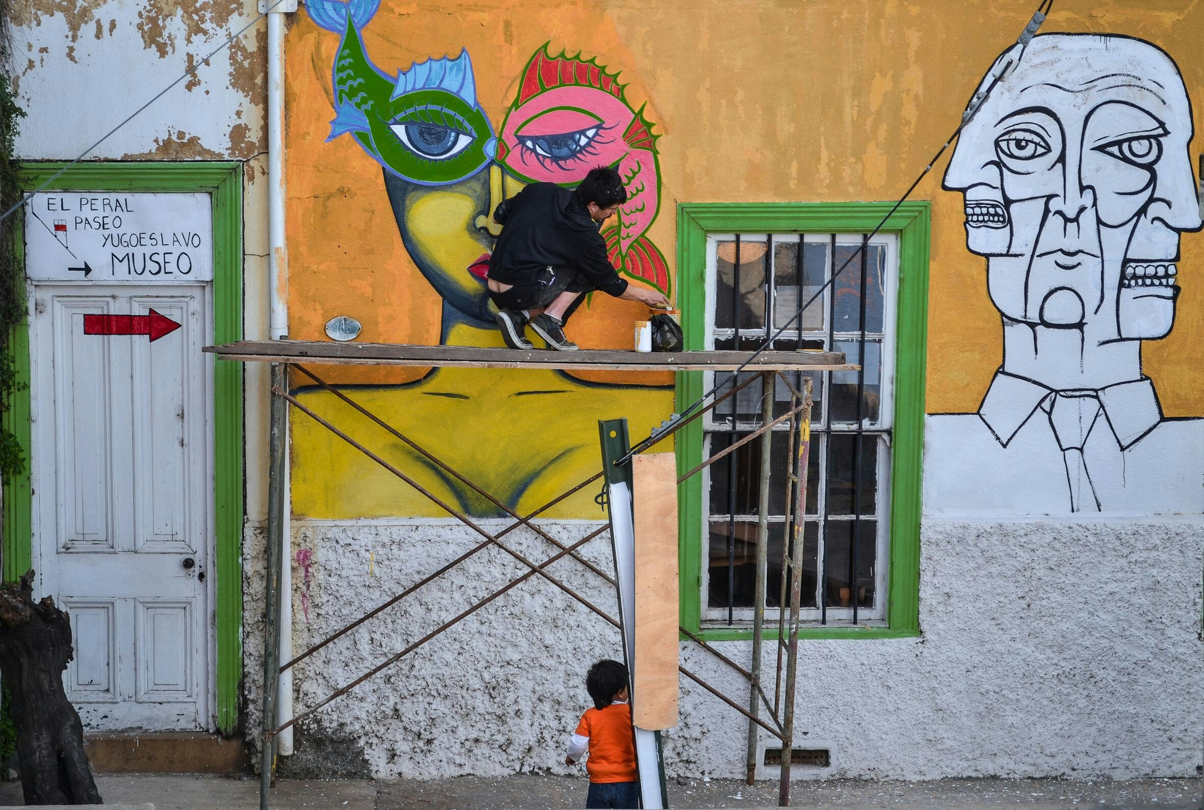 A street artist crouches on scaffolding painting a colourful mural on a building wall in Valparaiso, Chile, while a child watches.