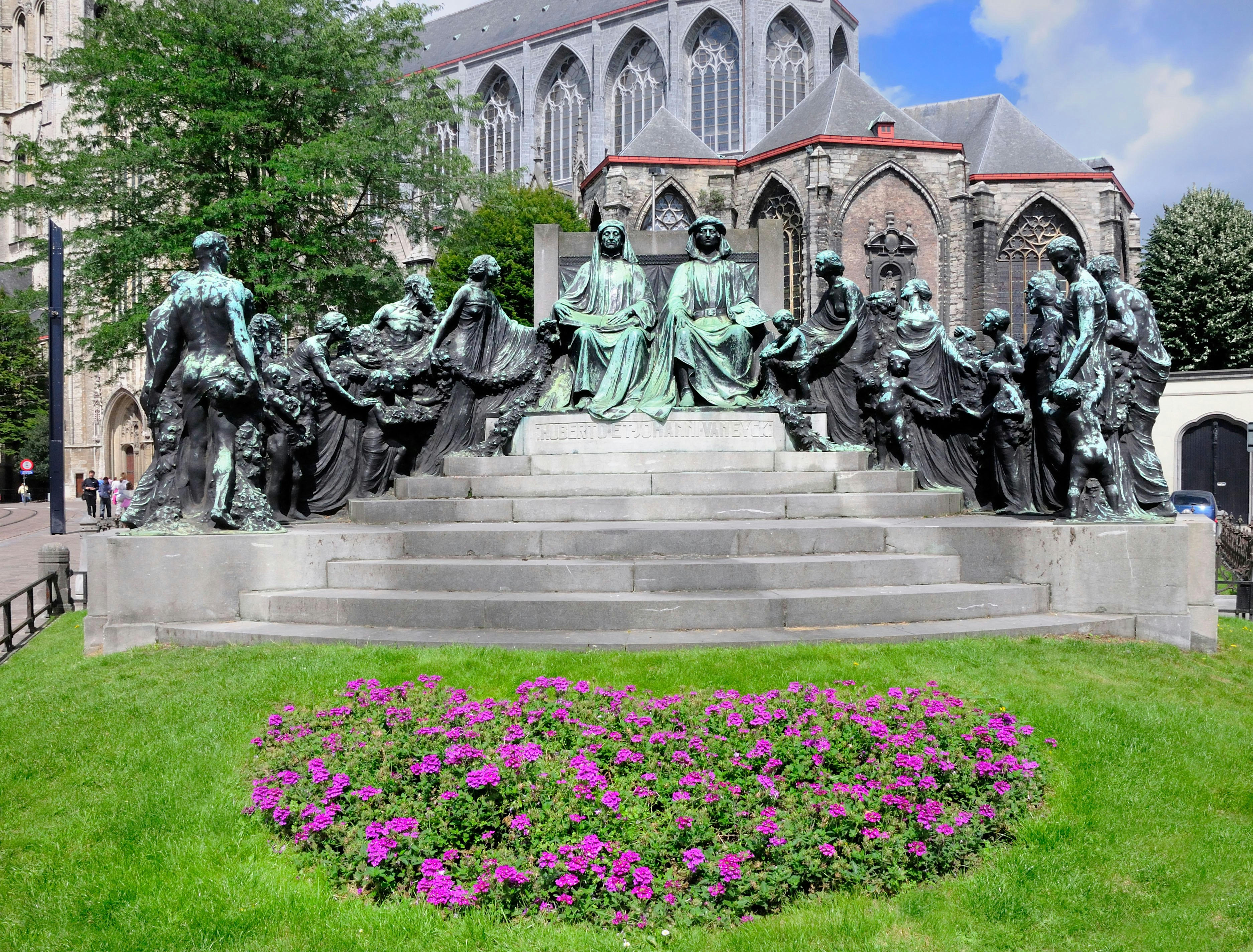 A statue of the Van Eyck brothers in Ghent, depicting the two artists sat side by side atop stone steps surrounded by statues of other people, with purple flowers in the foreground.