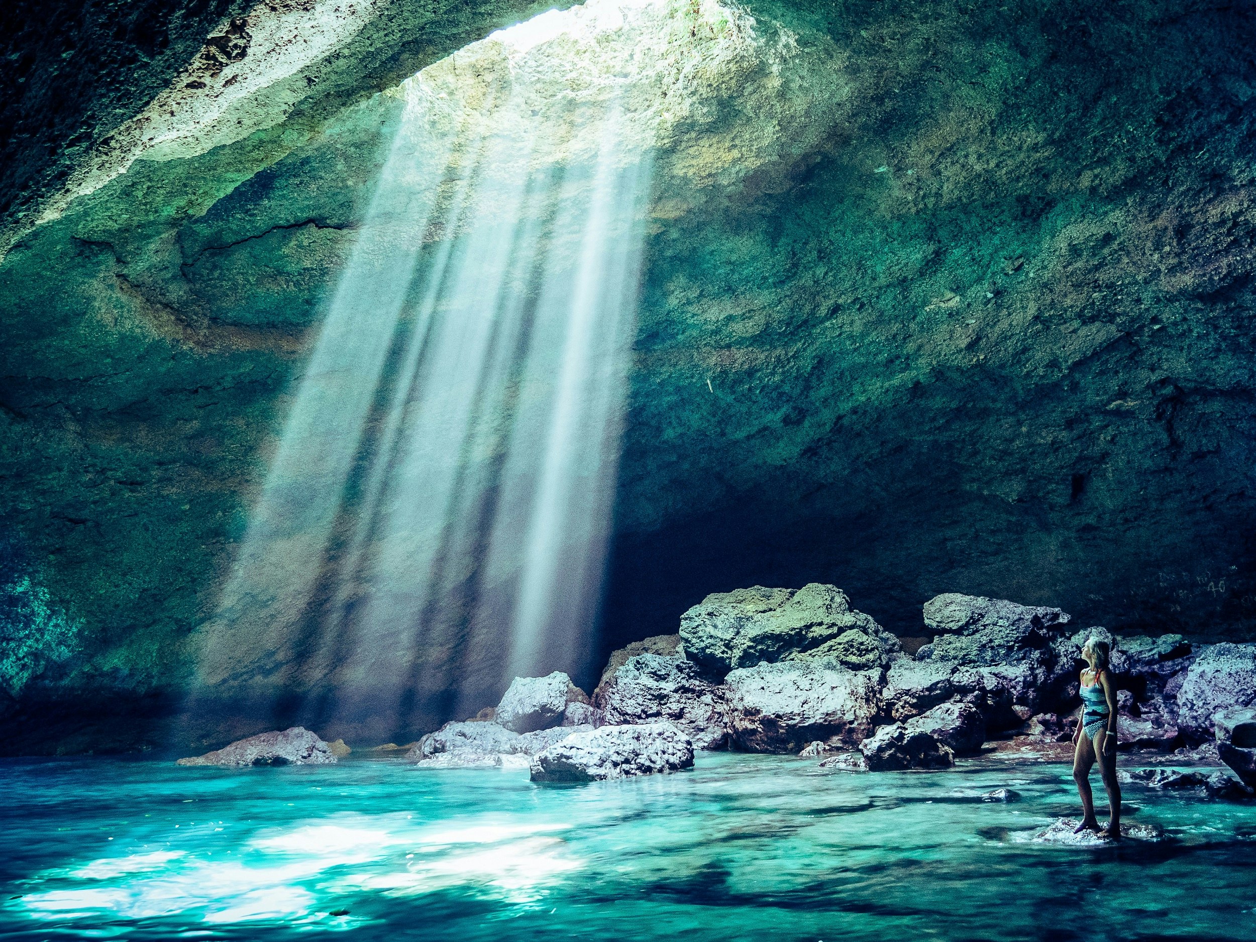 The author stands in a huge cave on a rock that is at the water's surface and looks up to brilliant shafts of light raining down from a hole in the top of the cave; the light that is hitting the water is causing it to glow a turquoise colour. The whole scene is bathed in a green hue.