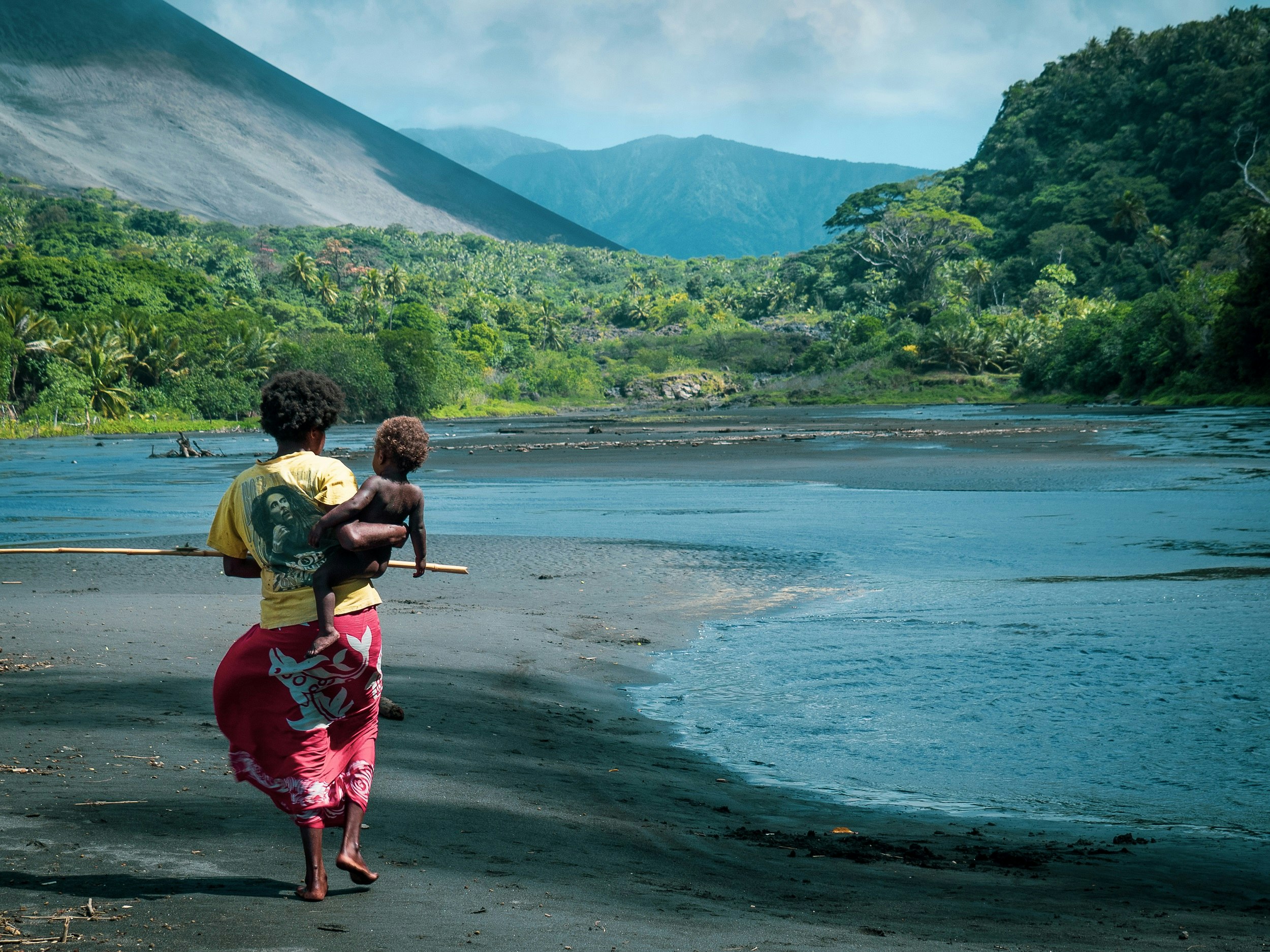 A local woman wearing a red sarong and yellow Bob Marley t-shirt carries a naked child in her arms as she walks long the river's edge on black sand; in the background is the slope of Mt Yasur and a tropical jungle at its base.
